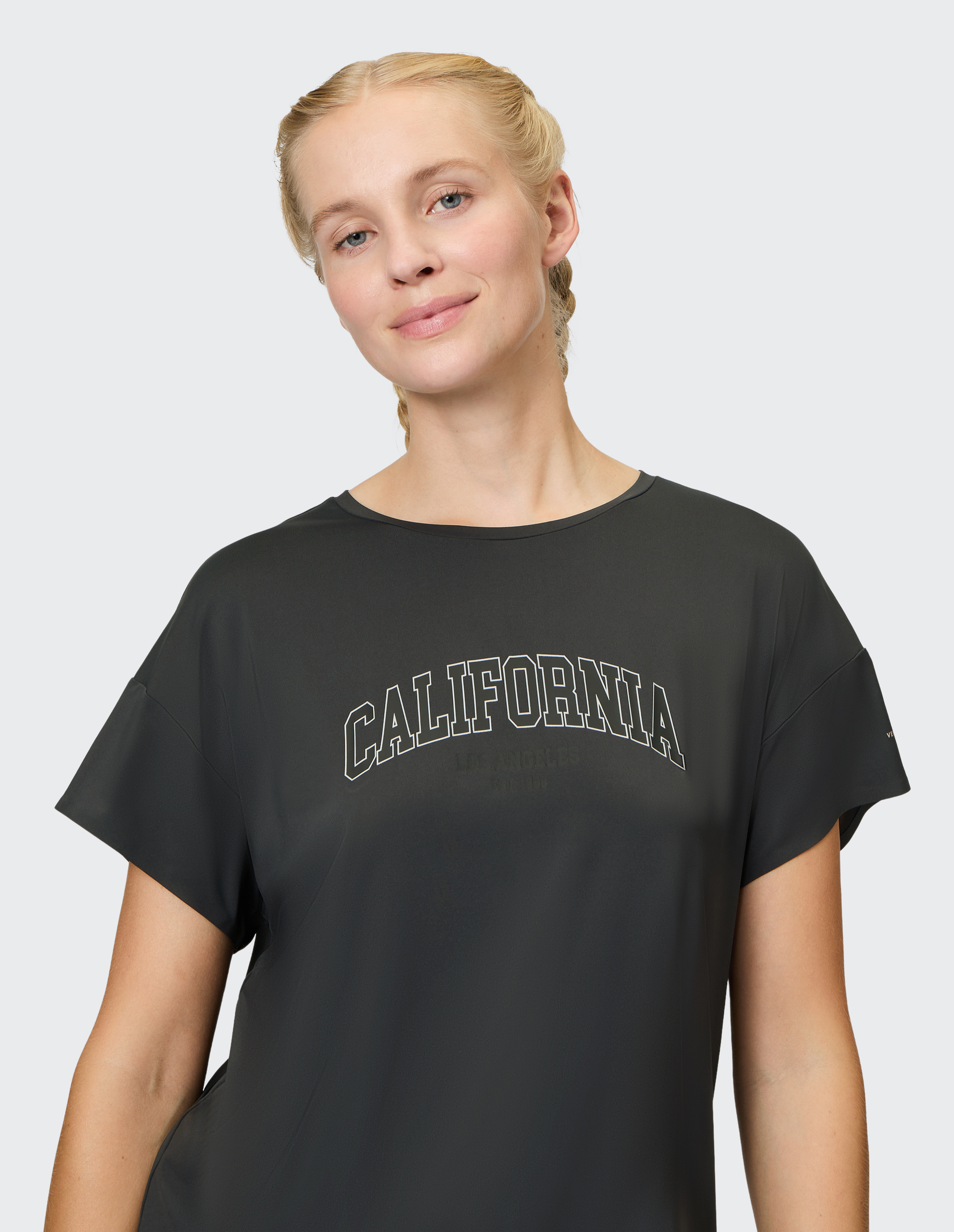 Eine lächelnde Person mit zwei geflochtenen Zöpfen trägt ein schwarzes T-Shirt mit der Aufschrift "California" auf der Vorderseite.