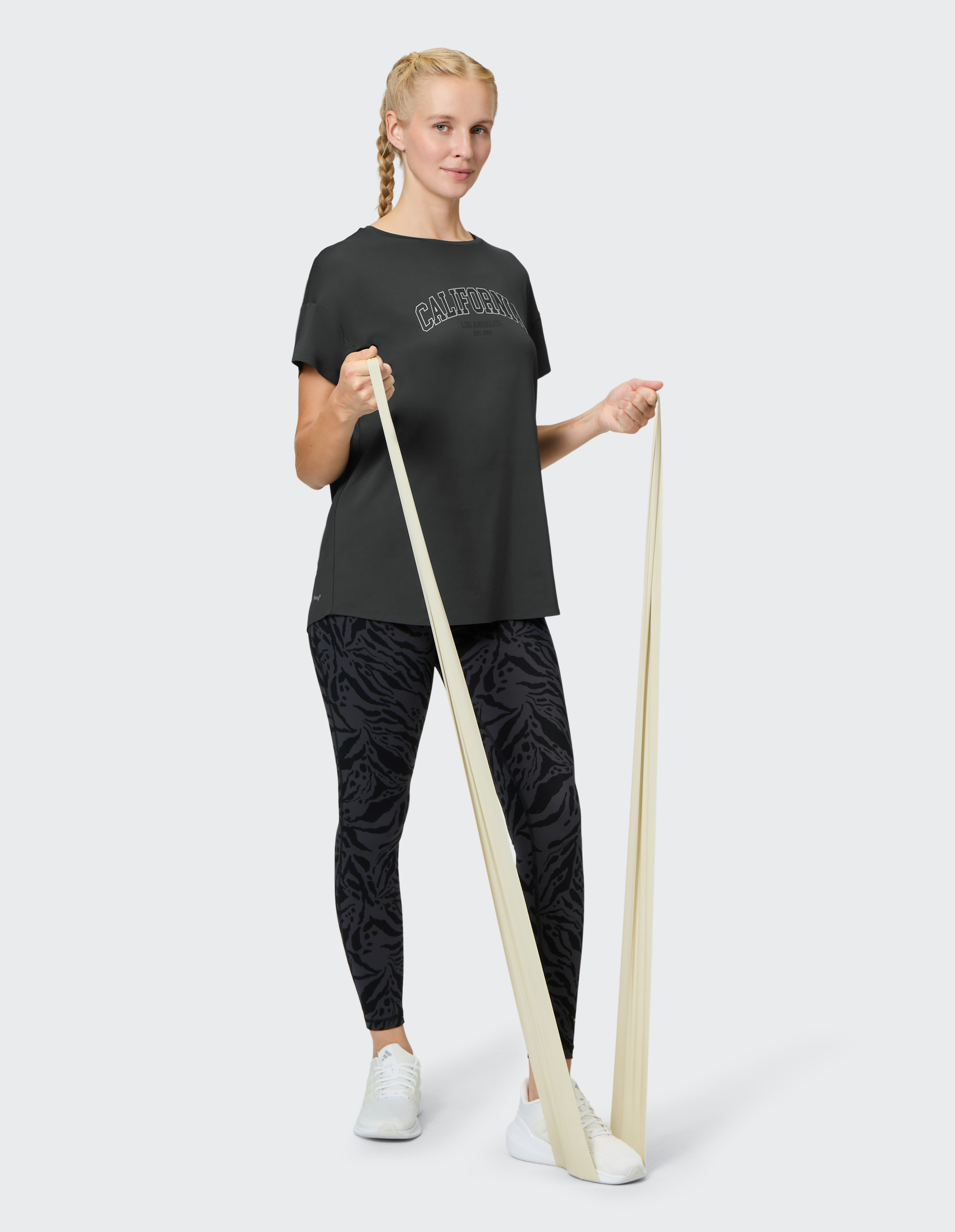 Frau mit geflochtenem Haar in schwarzem T-Shirt und gemusterten Leggings, trainiert mit Theraband um ihre Füße gewickelt.