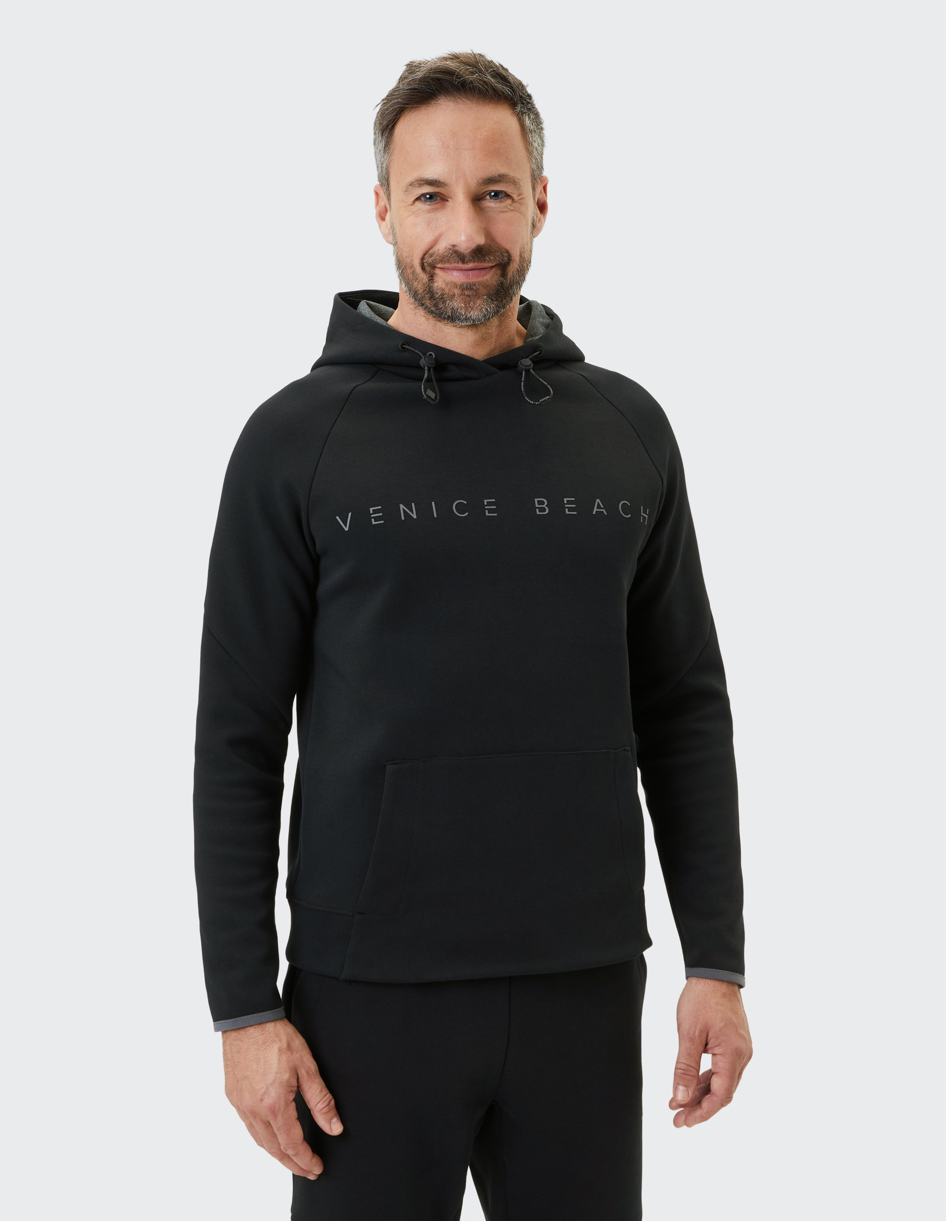 Ein lächelnder Mann trägt einen schwarzen Hoodie mit "Venice Beach"-Schriftzug, posiert vor hellem Hintergrund.