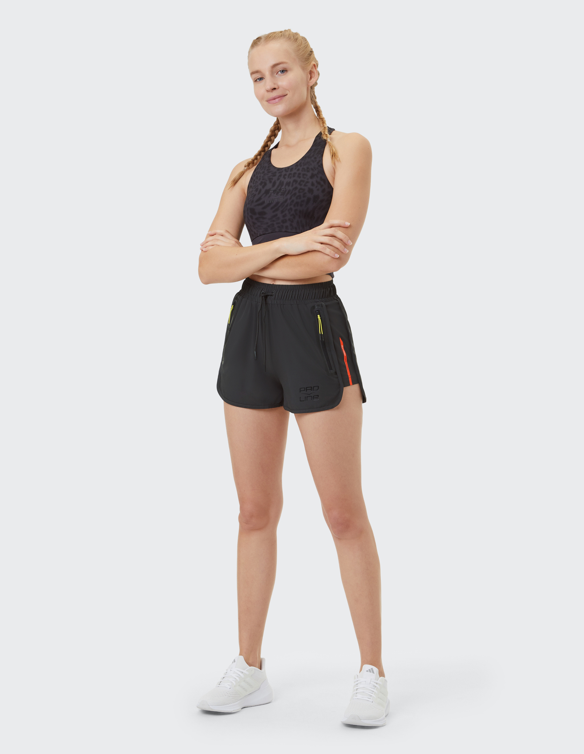 Eine Frau mit geflochtenem Haar trägt ein schwarzes Sporttop und kurze schwarze Shorts, posiert lächelnd mit verschränkten Armen.