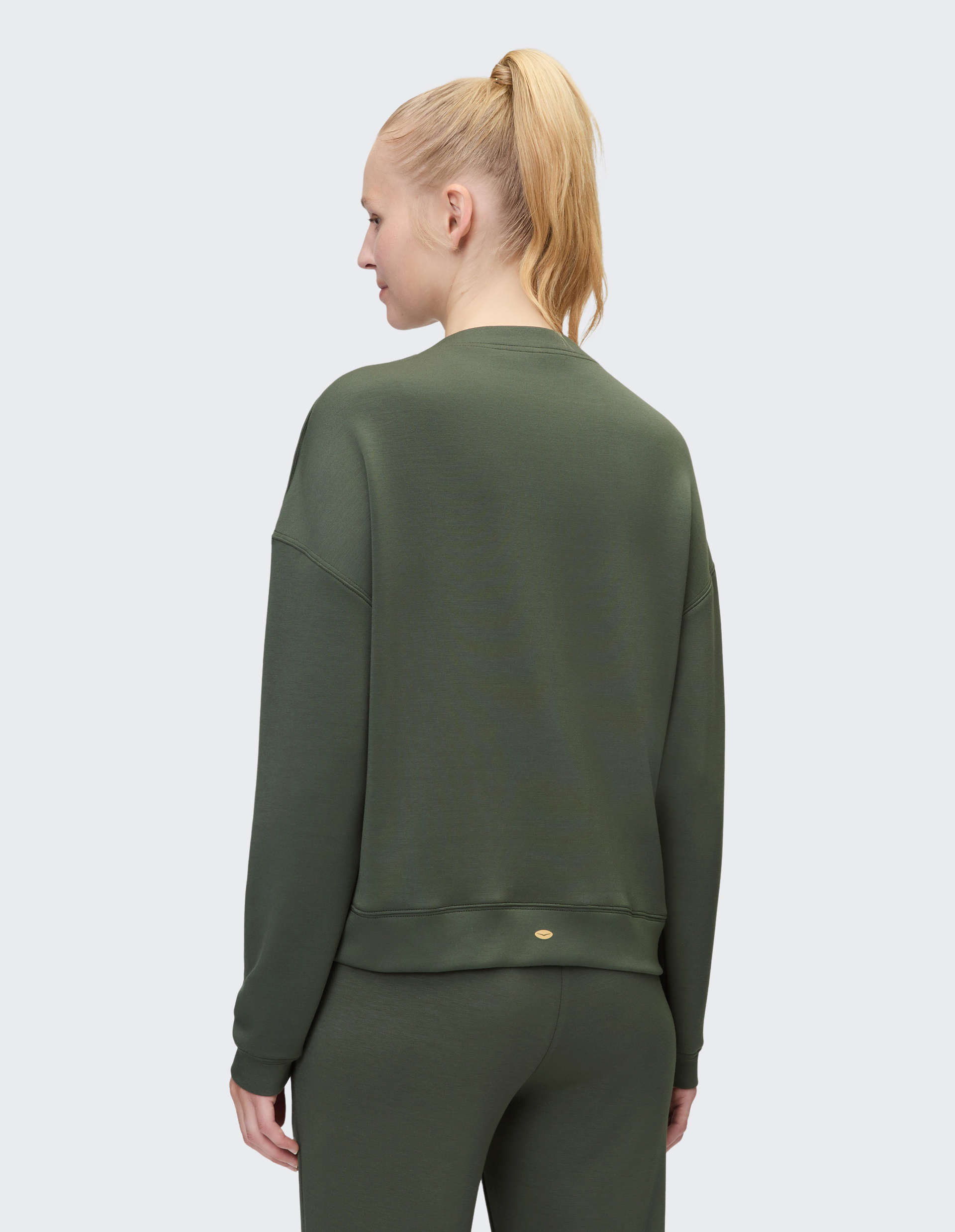 Person in olivgrünem, langärmeligem Sweatshirt und passender Hose, steht mit blondem Pferdeschwanz von hinten gesehen.