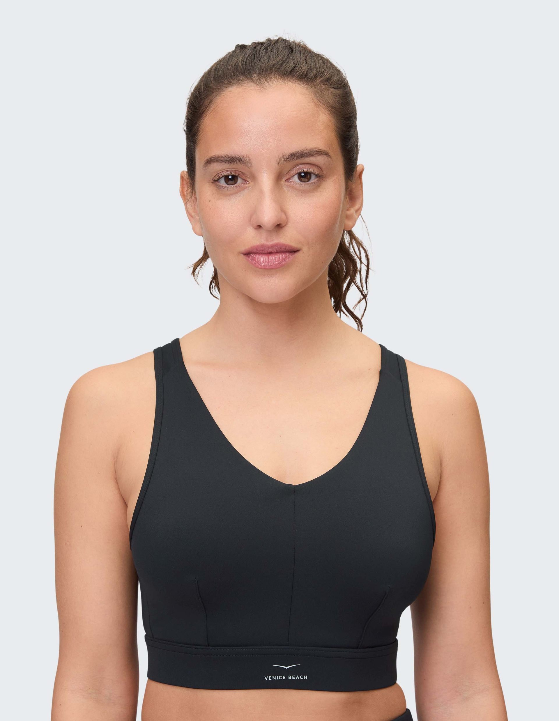 Frau mit brünettem Haar im Pferdeschwanz trägt einen schwarzen Sport-BH vor hellem Hintergrund und schaut freundlich in die Kamera.