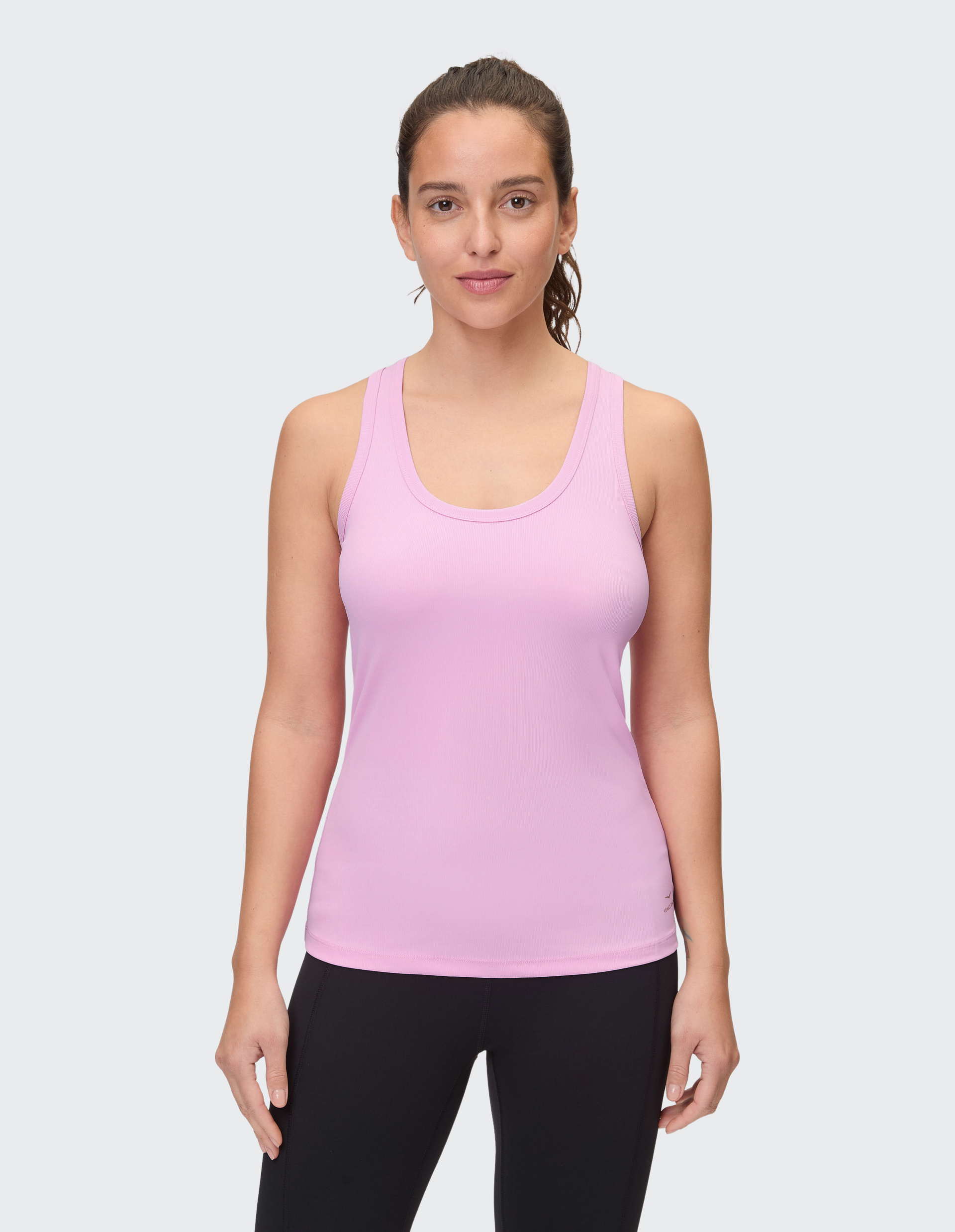 Frau mit seitlich gebundenem Haar trägt rosa Tanktop und schwarze Leggings, steht vor grauem Hintergrund und schaut freundlich.