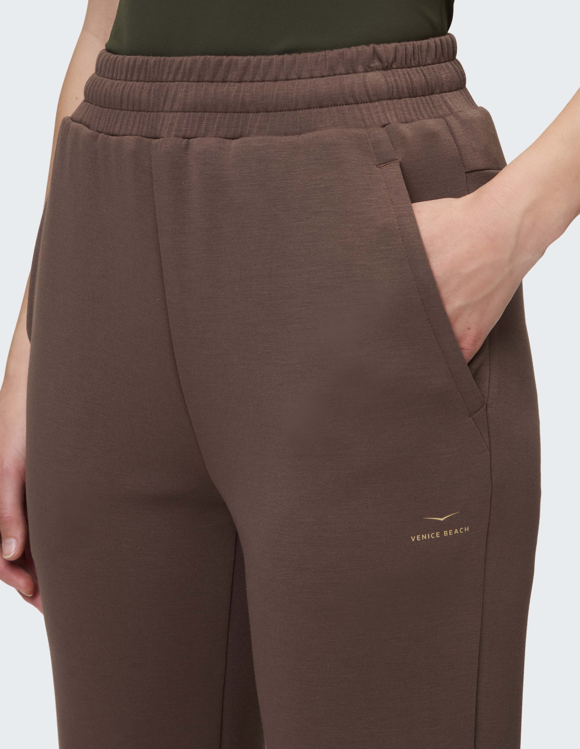Braune Jogginghose mit elastischem Bund und seitlicher Tasche; eine Hand ruht in der Tasche.