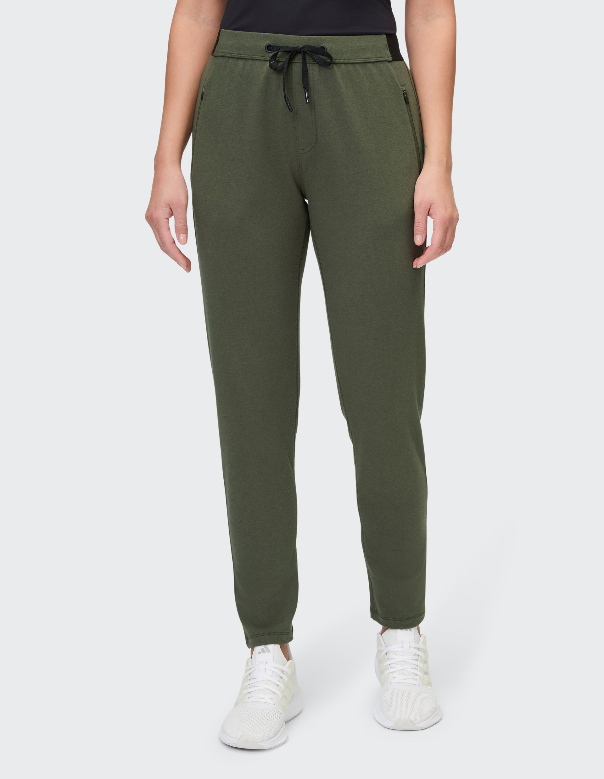 Frau trägt olivgrüne Jogginghose mit Kordelzug und weißen Sneakers, kombiniert mit einem schwarzen Shirt, vor weißem Hintergrund.