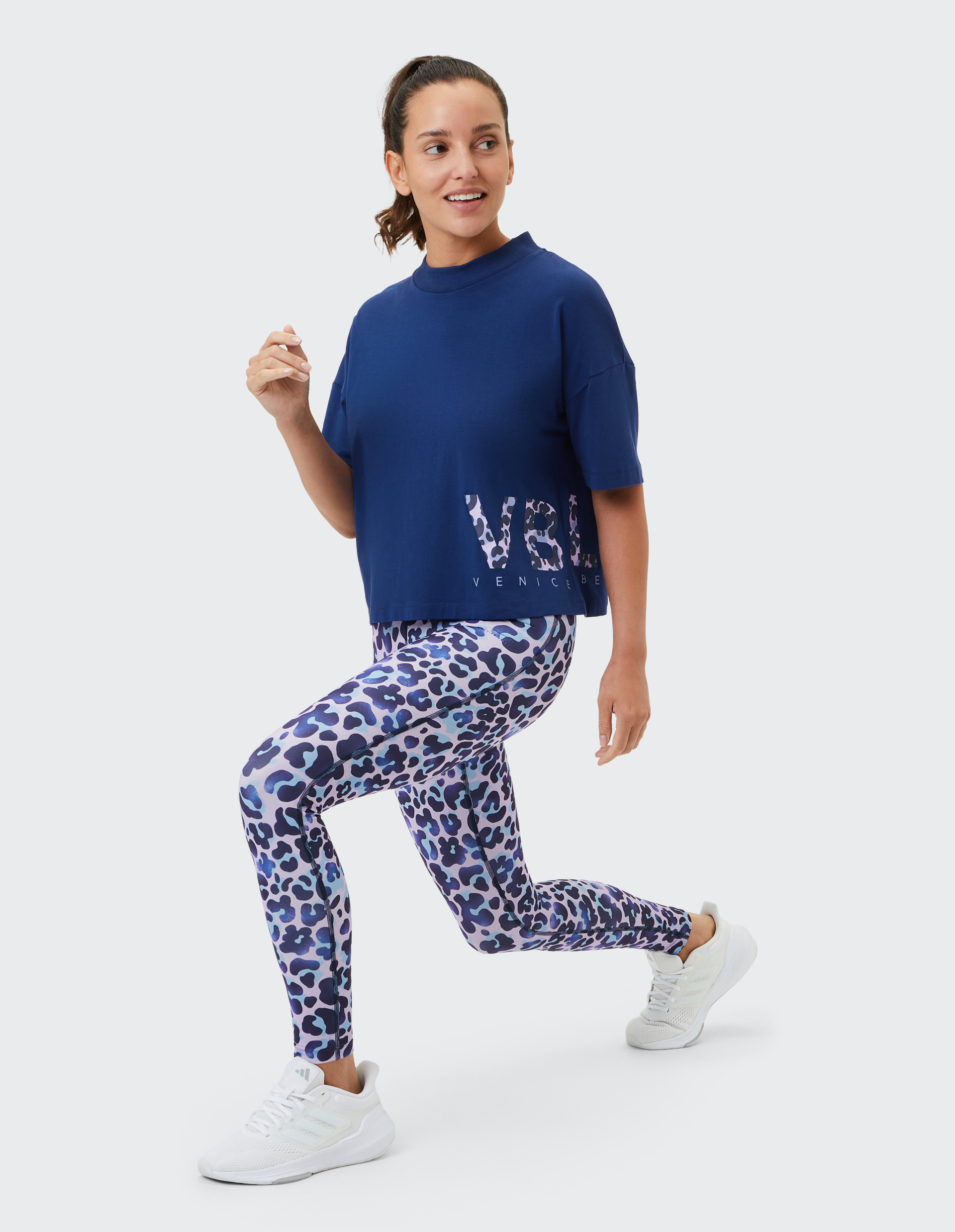 Frau im blauen Shirt und lila Leoparden-Leggings macht Ausfallschritt, trägt weiße Sneakers und hat Haare locker zum Zopf gebunden.