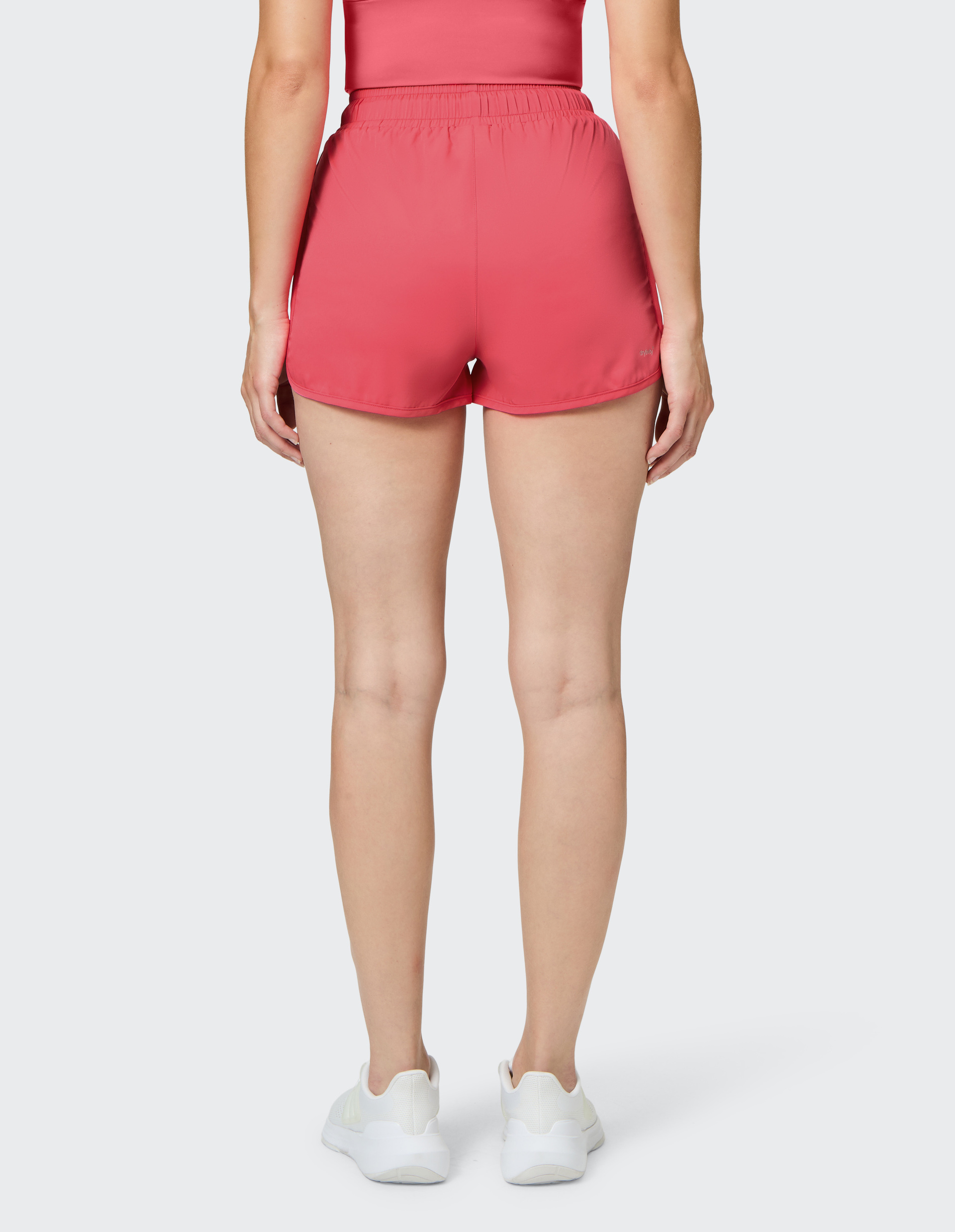Person in roten Shorts und passendem Top, aufgenommen von hinten, trägt weiße Turnschuhe auf hellem Hintergrund.