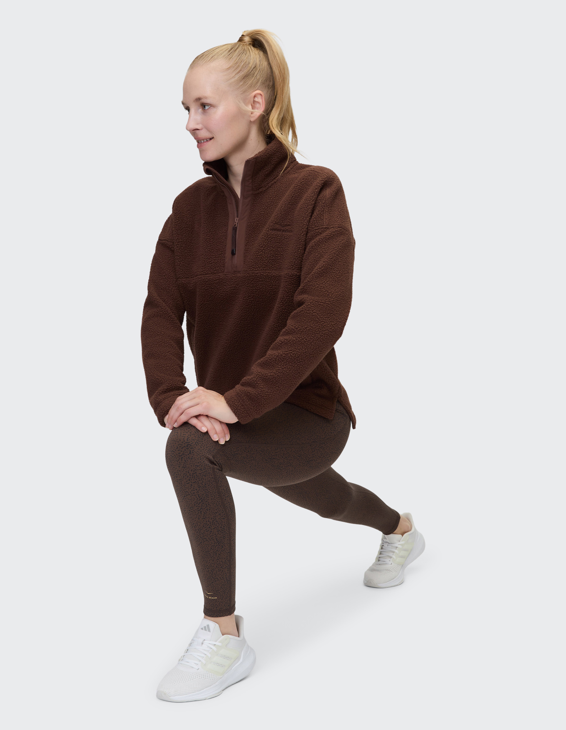 Eine Person trägt einen braunen Fleecepullover, gemusterte Leggings in Braun und weiße Turnschuhe vor einem grauen Hintergrund.
