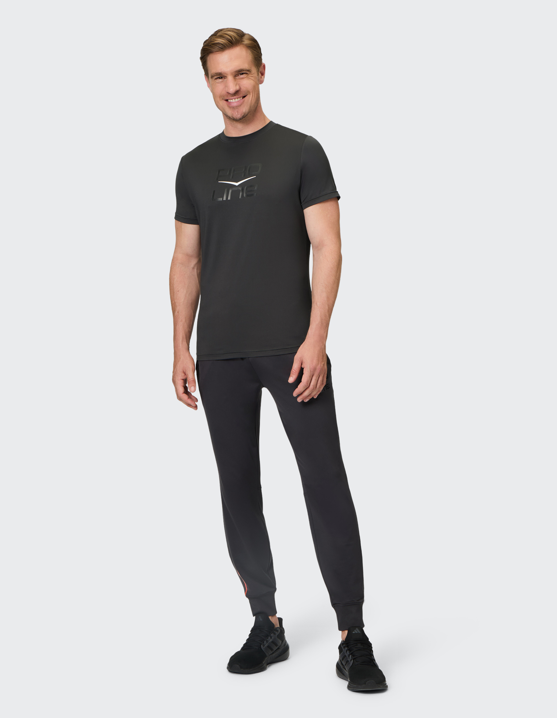 Ein lächelnder Mann trägt ein schwarzes T-Shirt und Jogginghose, steht aufrecht und schaut direkt in die Kamera, vor hellem Hintergrund.