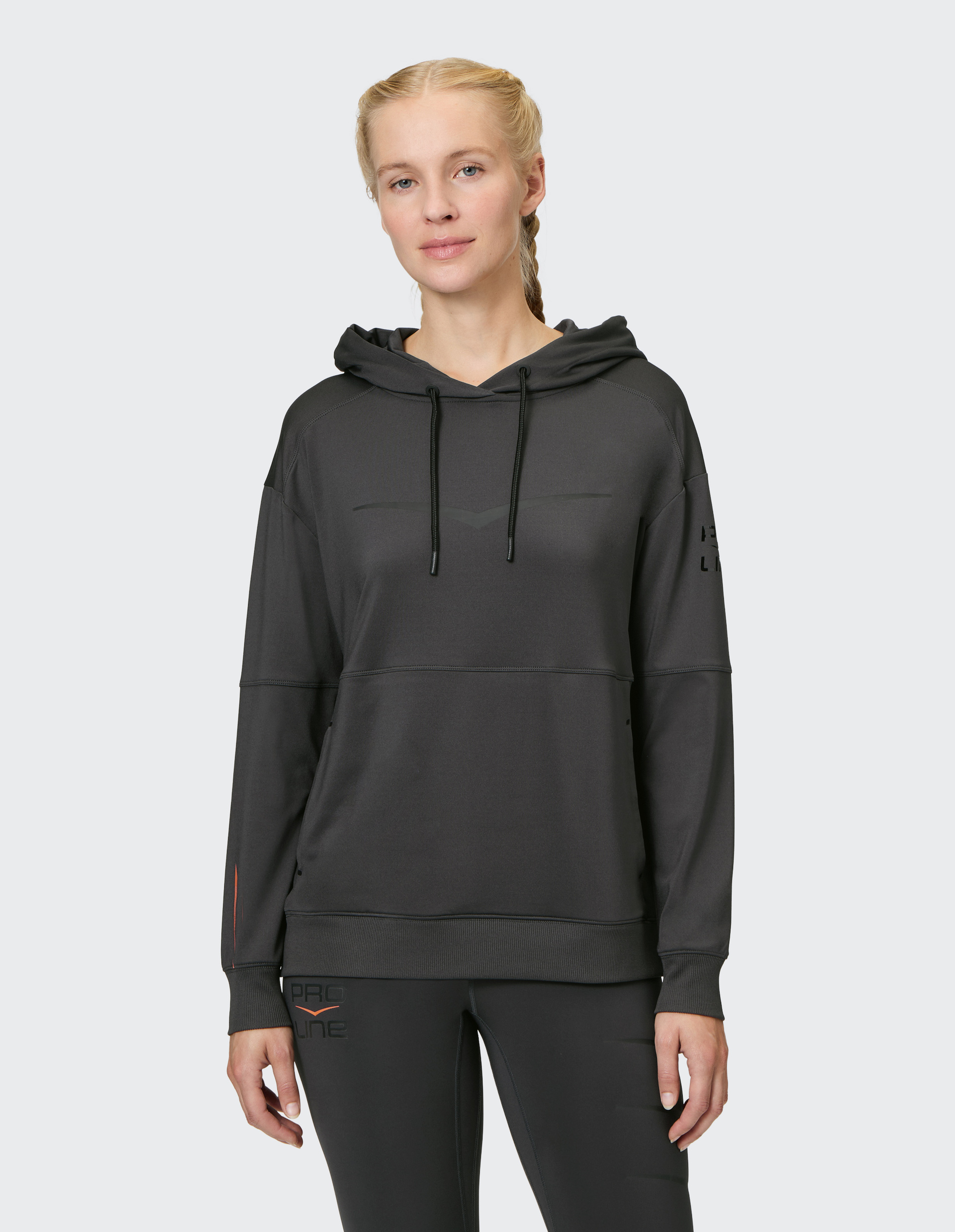 Eine Frau mit geflochtenem Haar trägt einen schwarzen Hoodie mit Kapuze und schwarzer Sportleggings vor einem neutralen Hintergrund.
