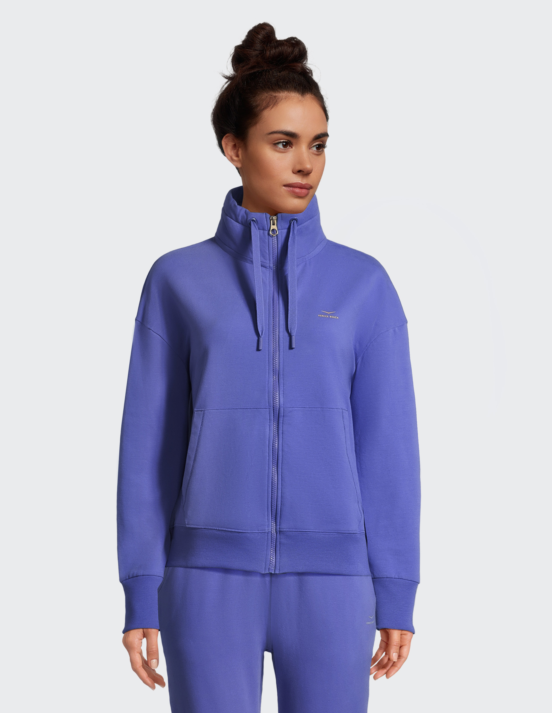 Frau trägt einen lilafarbenen Hoodie mit hohem Kragen und Reißverschluss, ihre Haare sind hochgesteckt, vor neutralem Hintergrund.