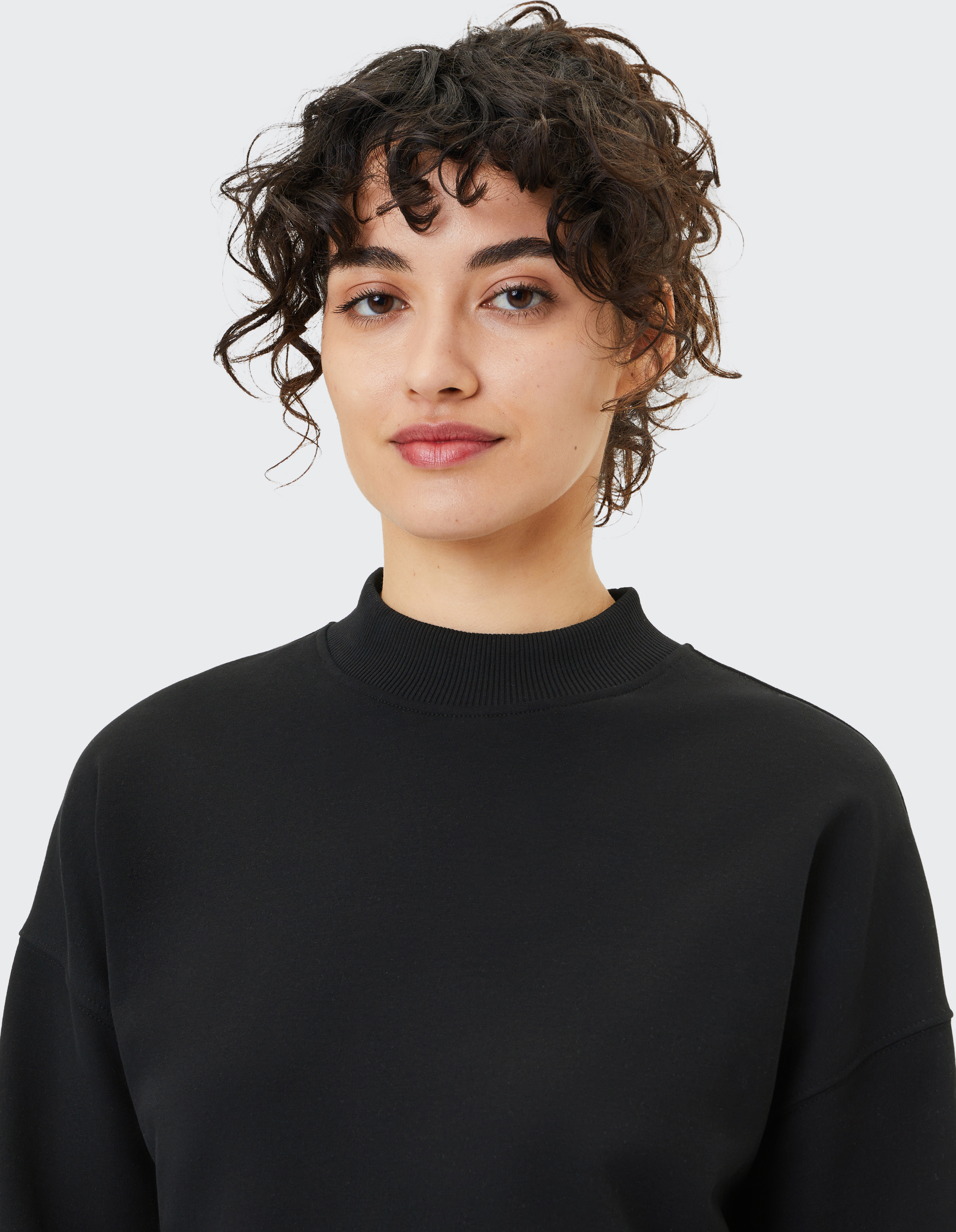 Eine Frau mit lockigem Haar und neutralem Gesichtsausdruck trägt einen schwarzen Pullover vor einem grauen Hintergrund.