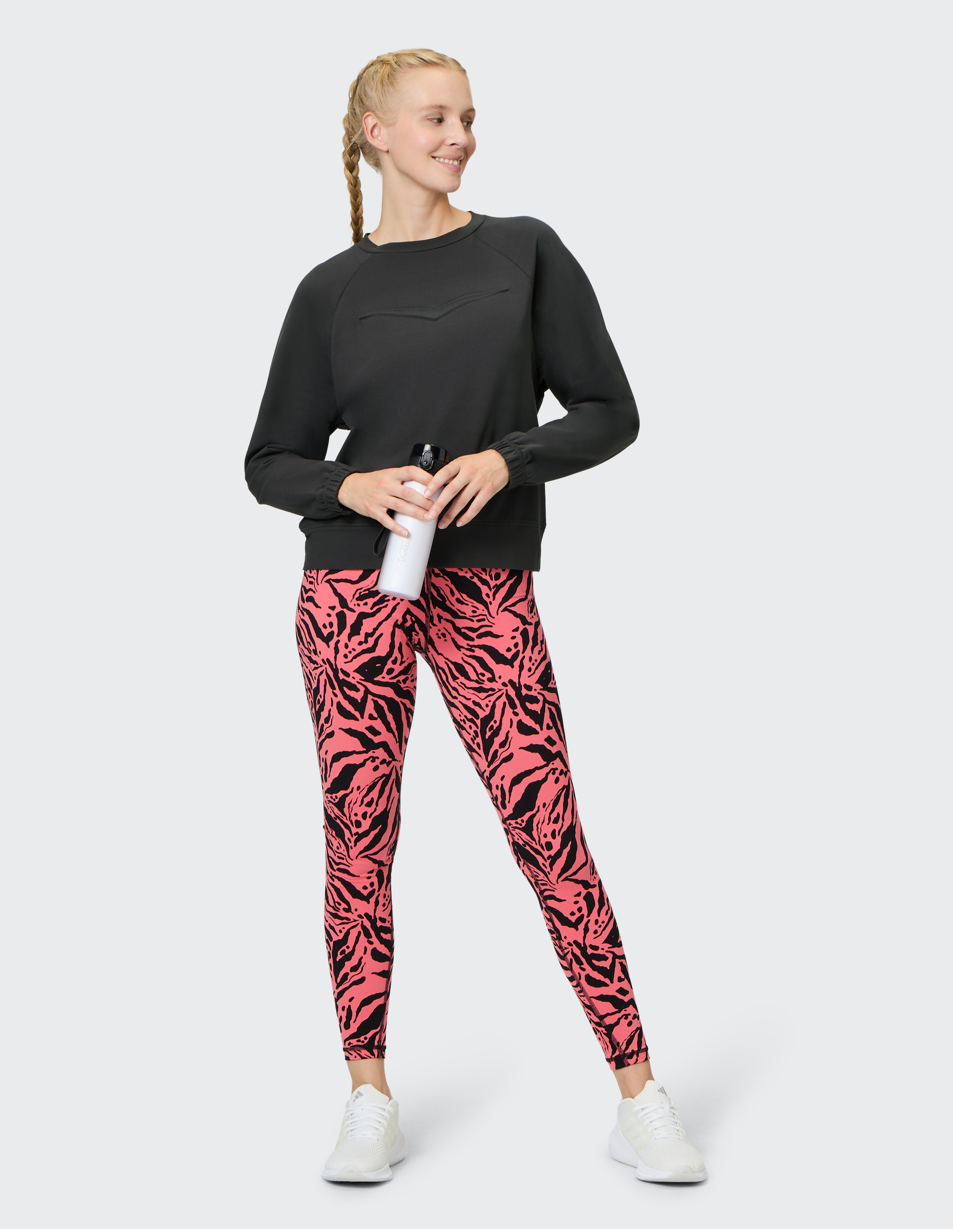 Frau in schwarzem Pullover und pinken, gemusterten Leggings hält eine Wasserflasche und lächelt, mit tragenden weißen Sneakers.