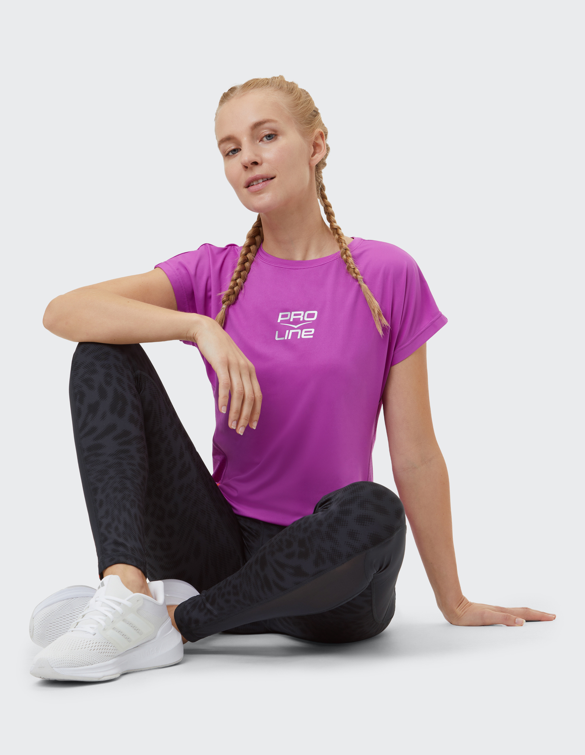 Frau in pinkem Sportshirt und schwarzen gemusterten Leggings sitzt entspannt mit weißen Sneakers auf hellem Boden, zwei Zöpfe frisur.