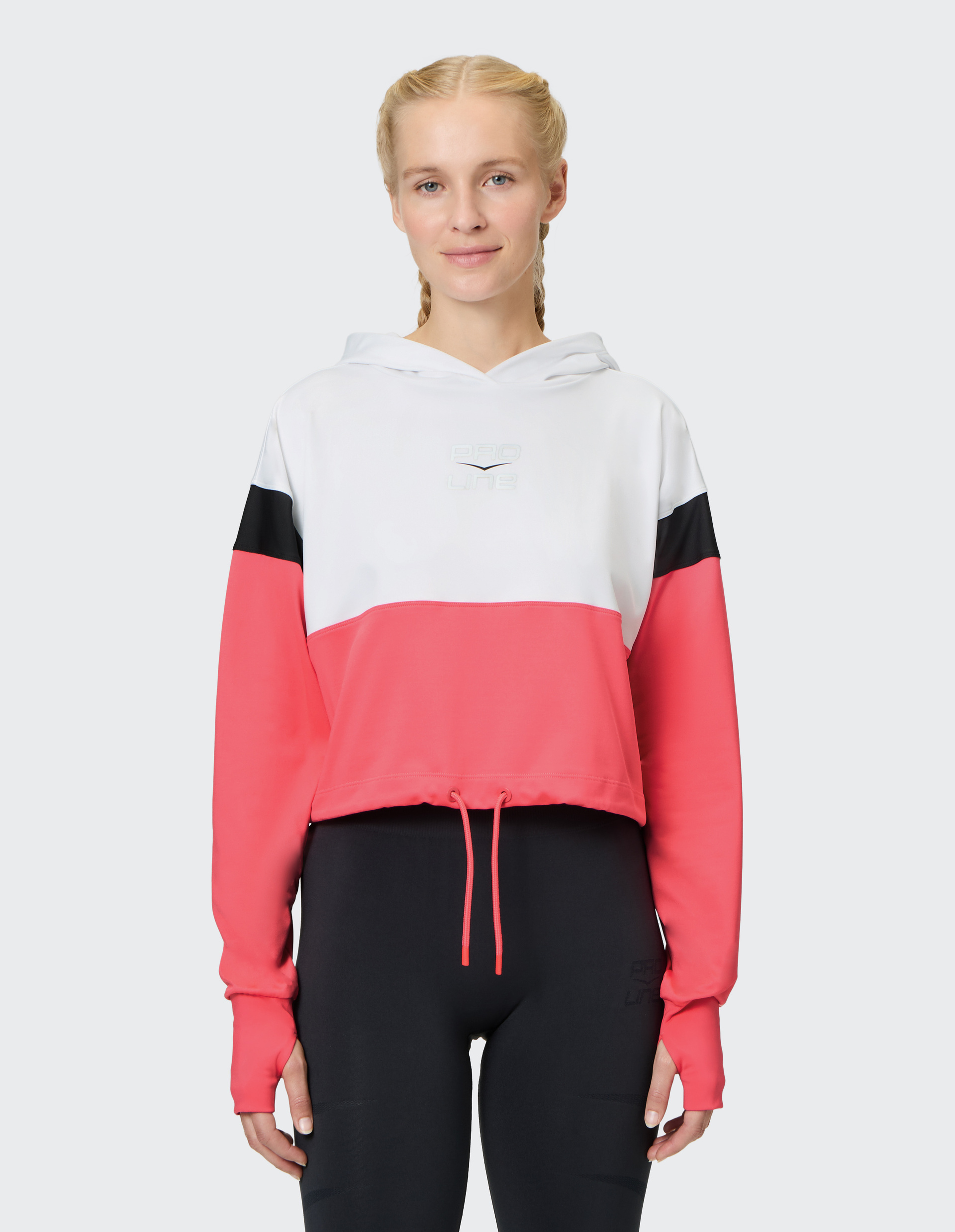 Frau mit geflochtenen Haaren trägt einen sportlichen Hoodie in Weiß, Pink und Schwarz sowie schwarze Leggings, auf hellem Hintergrund.