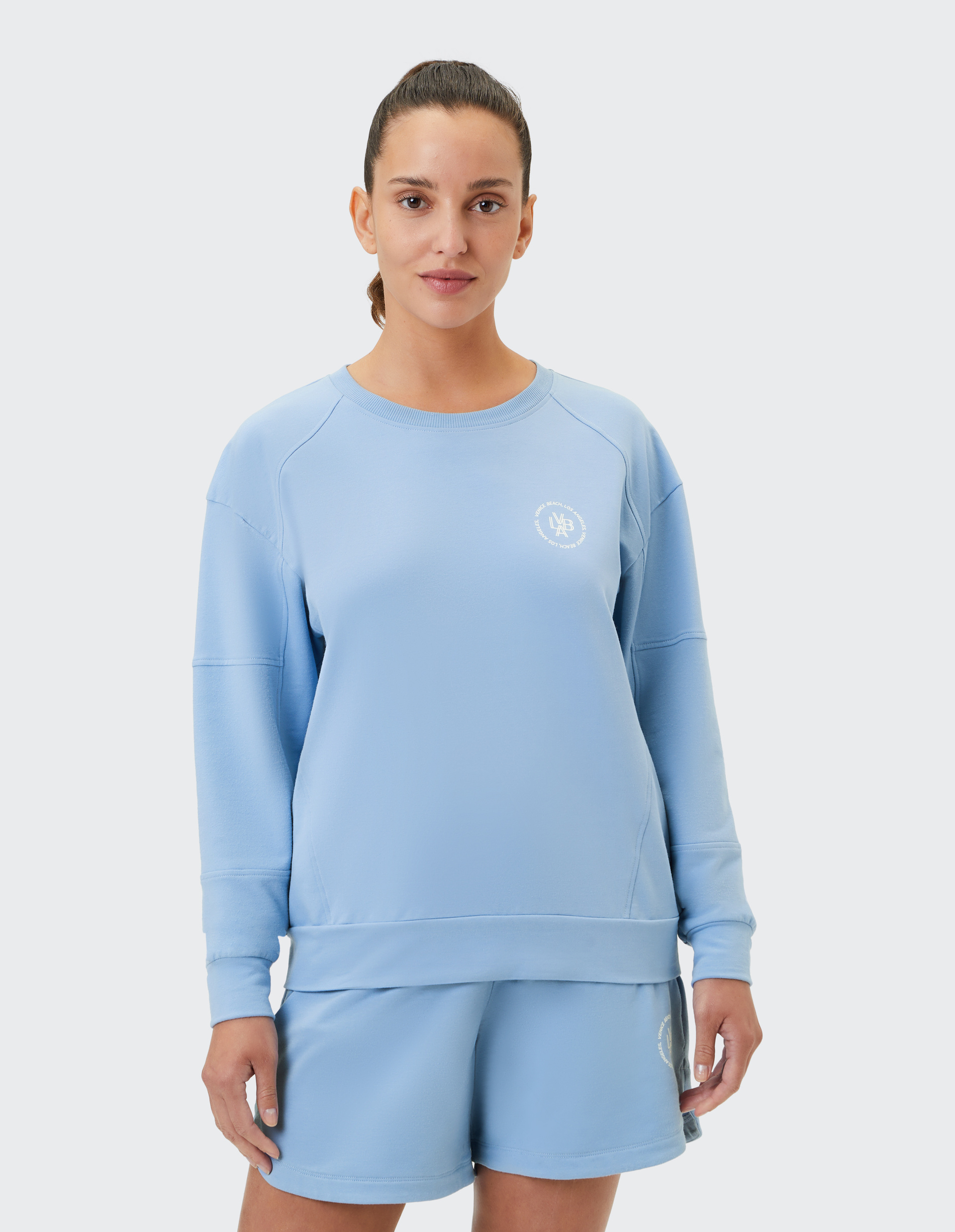 Eine lachende Person trägt einen hellblauen Sweater und passende Shorts vor einem hellgrauen Hintergrund.