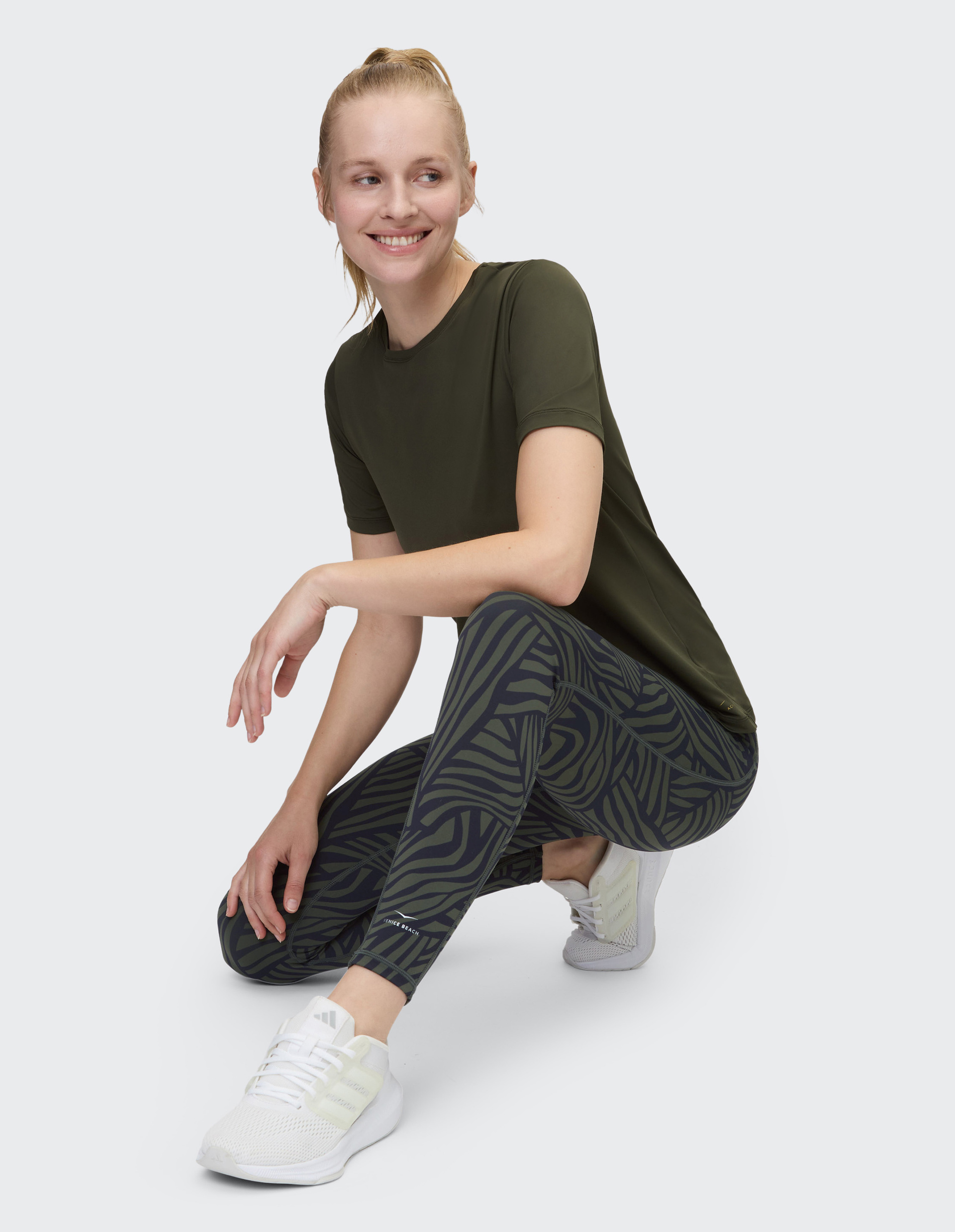 Person in olivgrünem Shirt und gemusterten Leggings steht lächelnd vor neutralem Hintergrund, trägt weiße Sneakers.