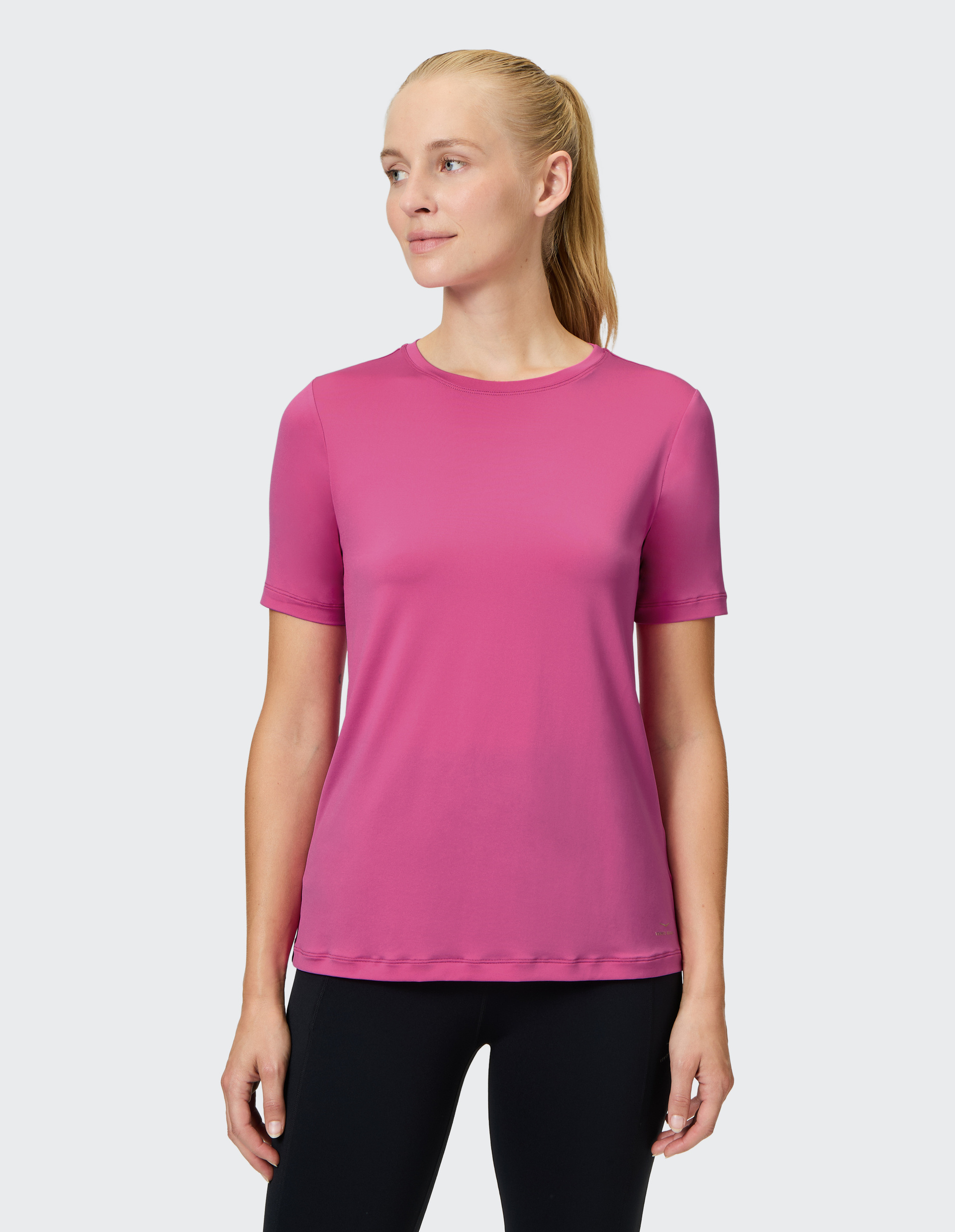 Eine Frau in einem rosa T-Shirt und schwarzen Leggings steht lächelnd vor einem neutralen Hintergrund und blickt nach links.