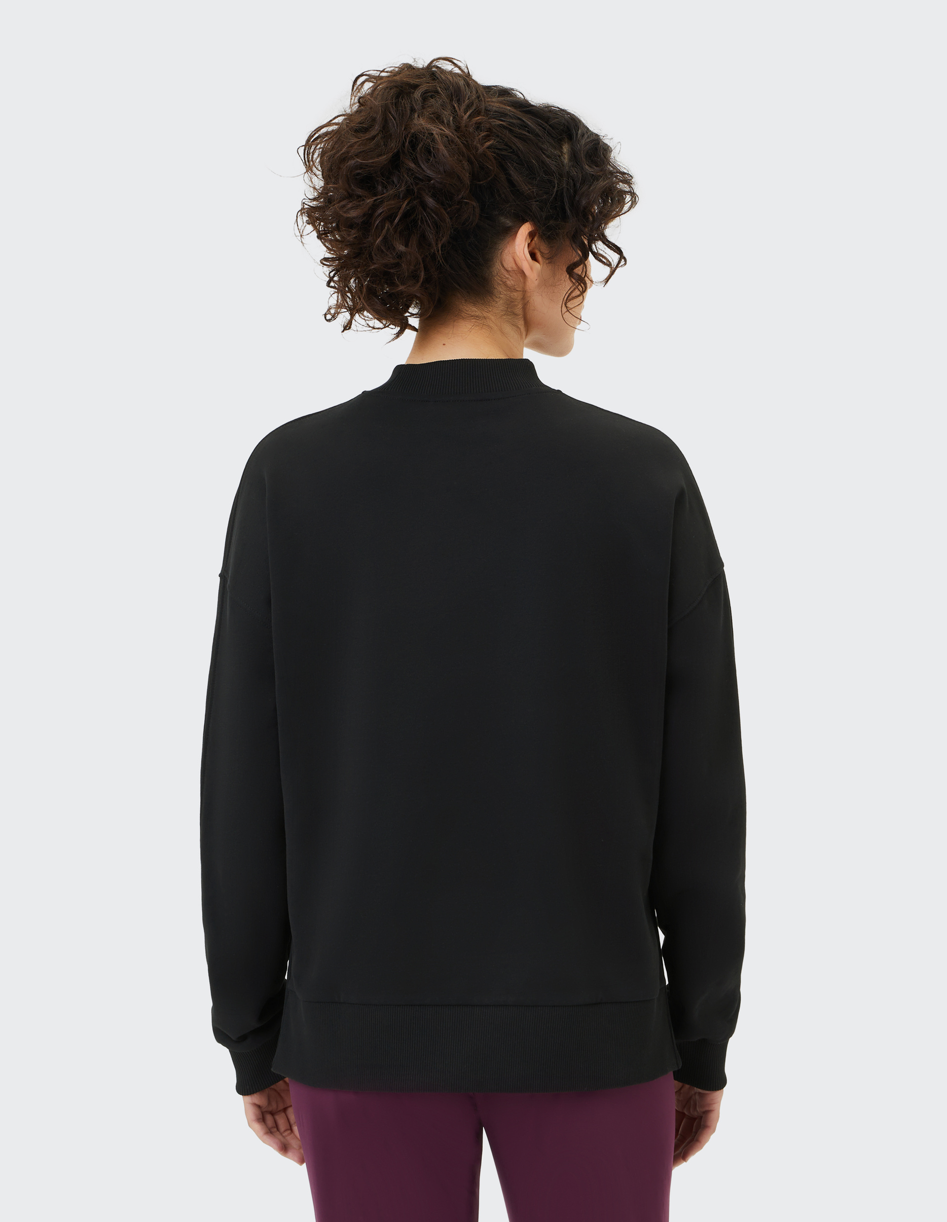 Person von hinten in schwarzem Sweatshirt und lila Hose, mit lockigem Haar, vor grauem Hintergrund stehend.