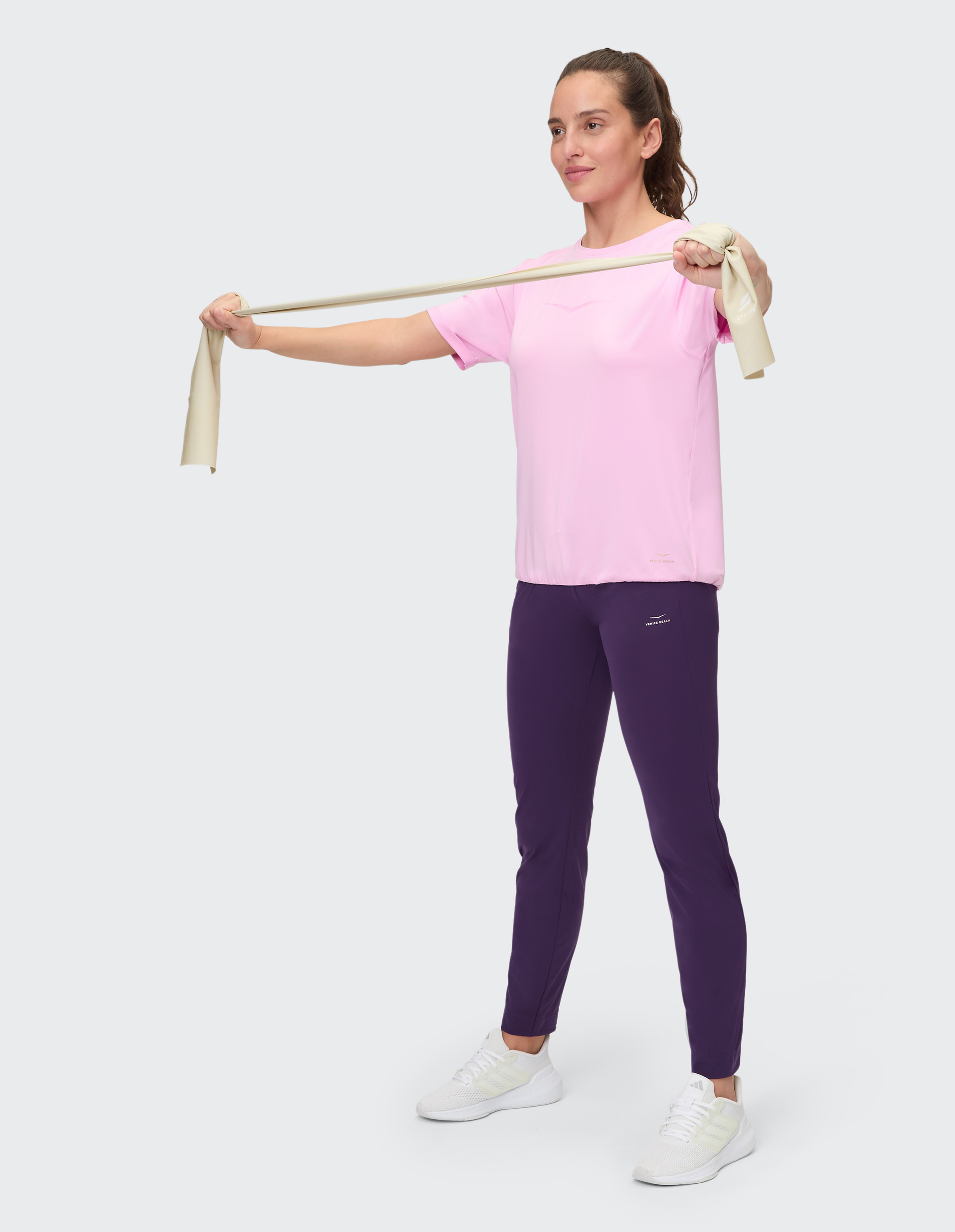 Eine Person trägt ein rosa T-Shirt und lila Jogginghosen, steht frontal vor einem hellgrauen Hintergrund und trägt weiße Schuhe.