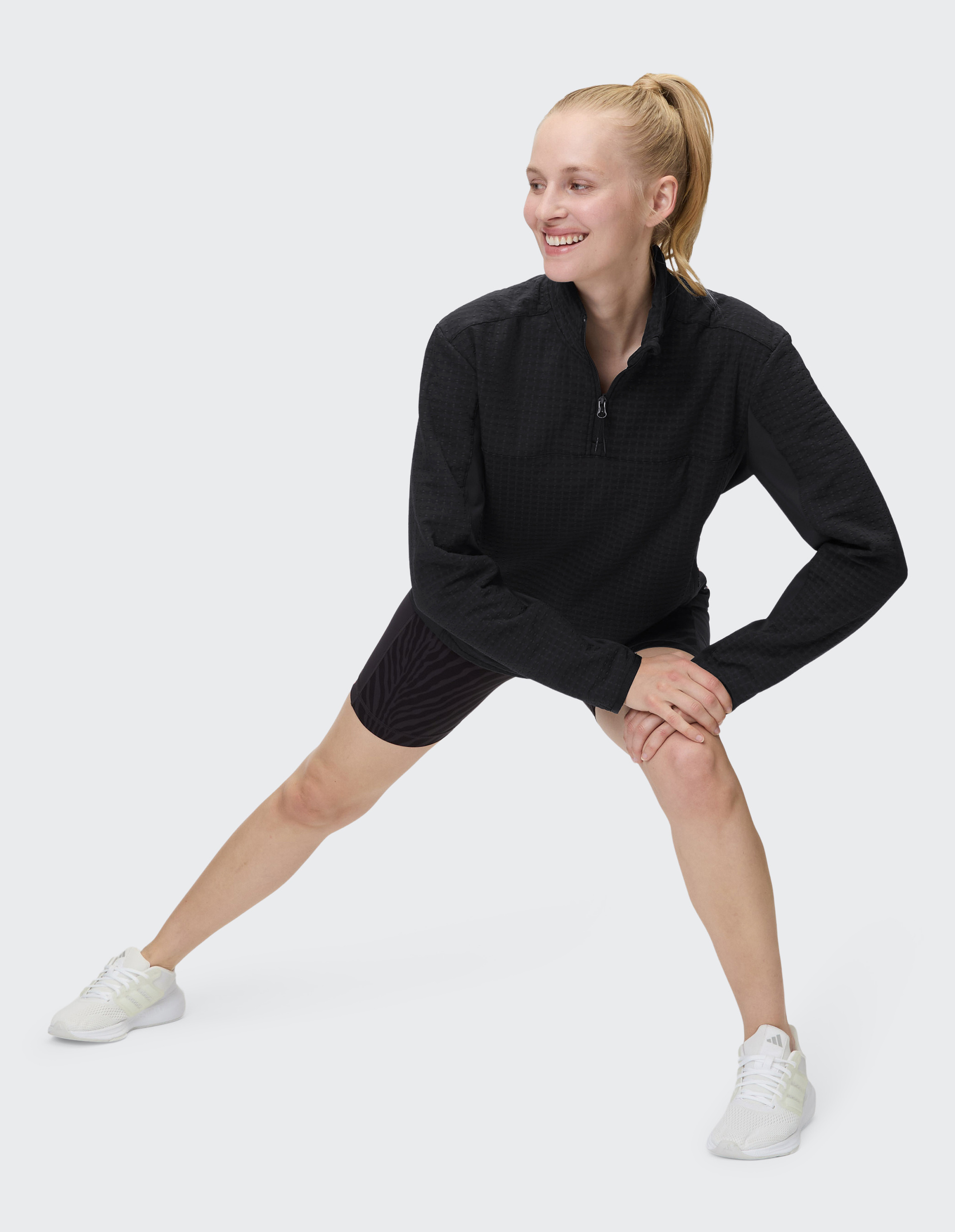 Frau mit blondem Pferdeschwanz dehnt in sportlichem Outfit, schwarzem Oberteil und weißen Schuhen lächelnd das linke Bein.