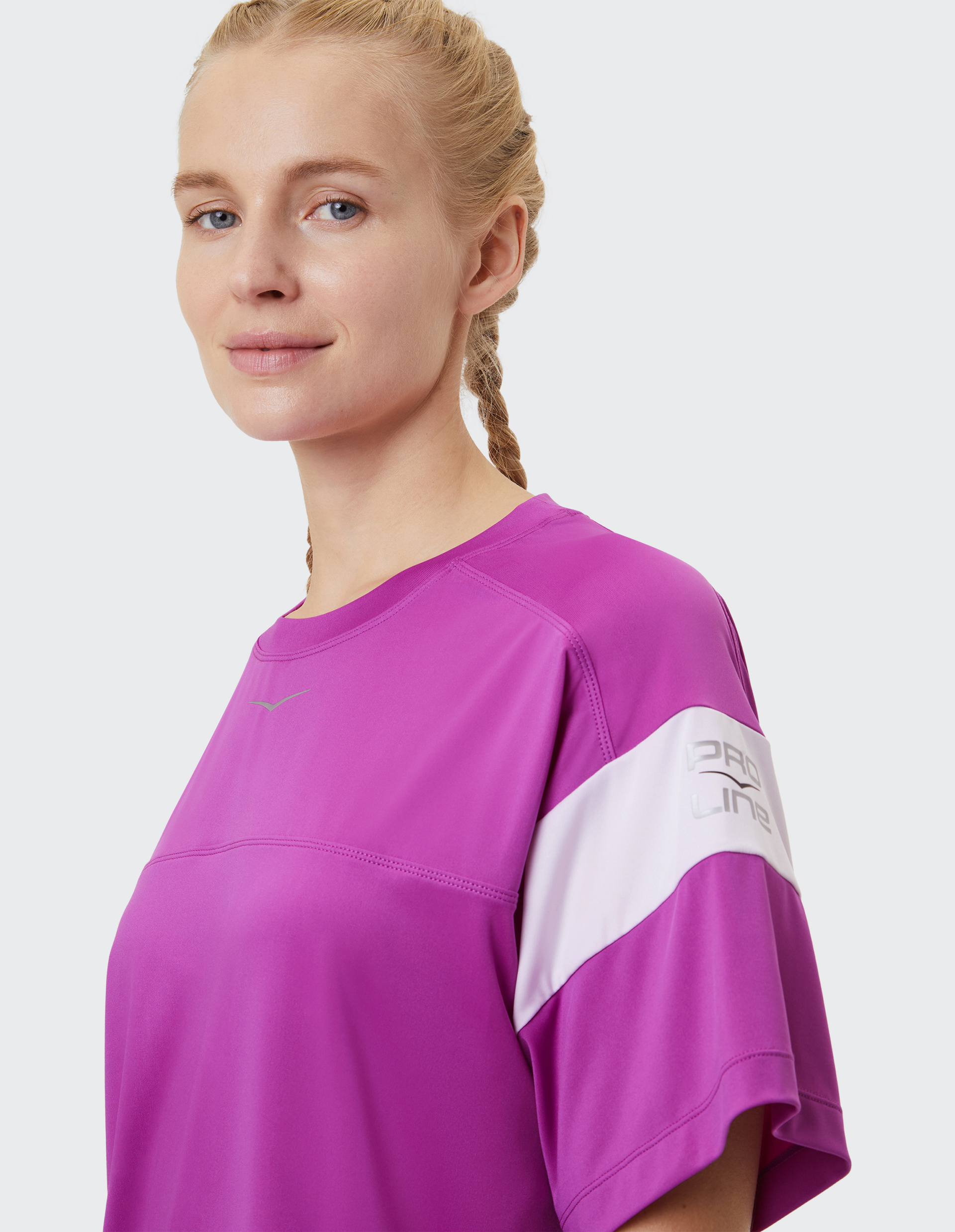 Frau mit blonden Zöpfen trägt ein lila T-Shirt mit weißem Streifen und Logo am Ärmel und lächelt leicht vor hellem Hintergrund.