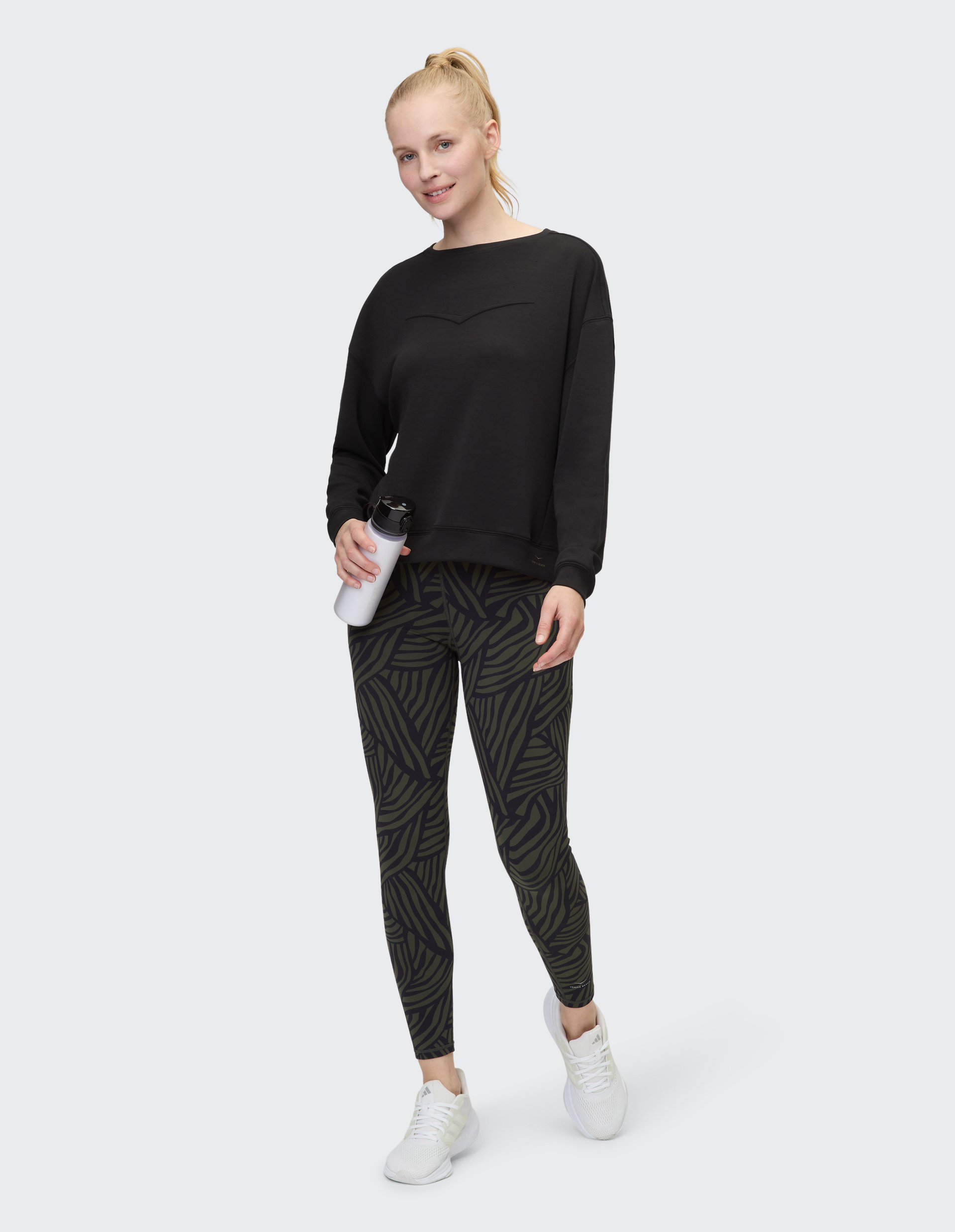 Person trägt schwarzen Pullover, grün-schwarze gemusterte Leggings und weiße Schuhe, steht auf grauem Hintergrund.