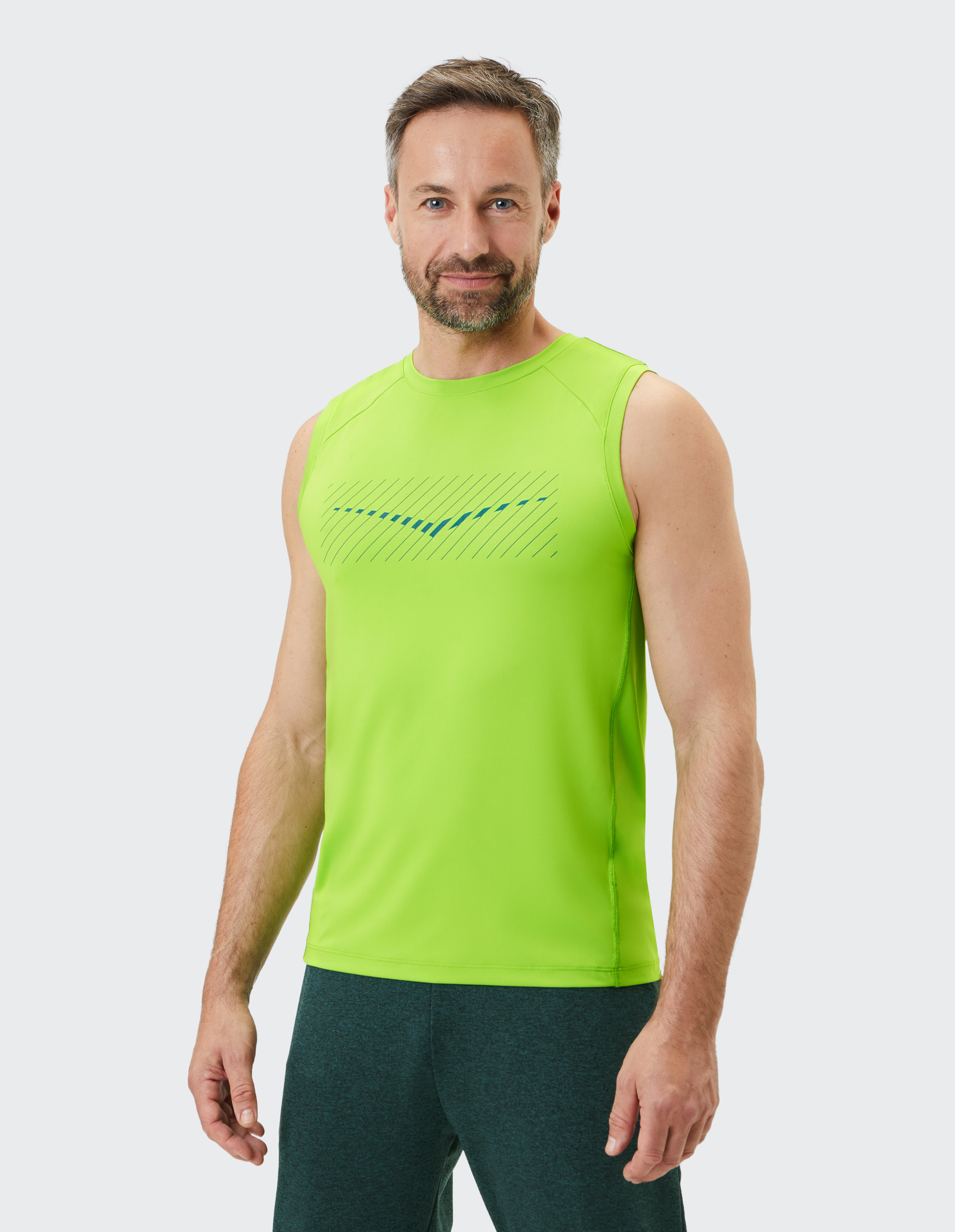 Ein Mann mit Bart trägt ein neon-grünes ärmelloses Shirt und dunkle Sporthosen, lächelnd vor einem hellen Hintergrund.