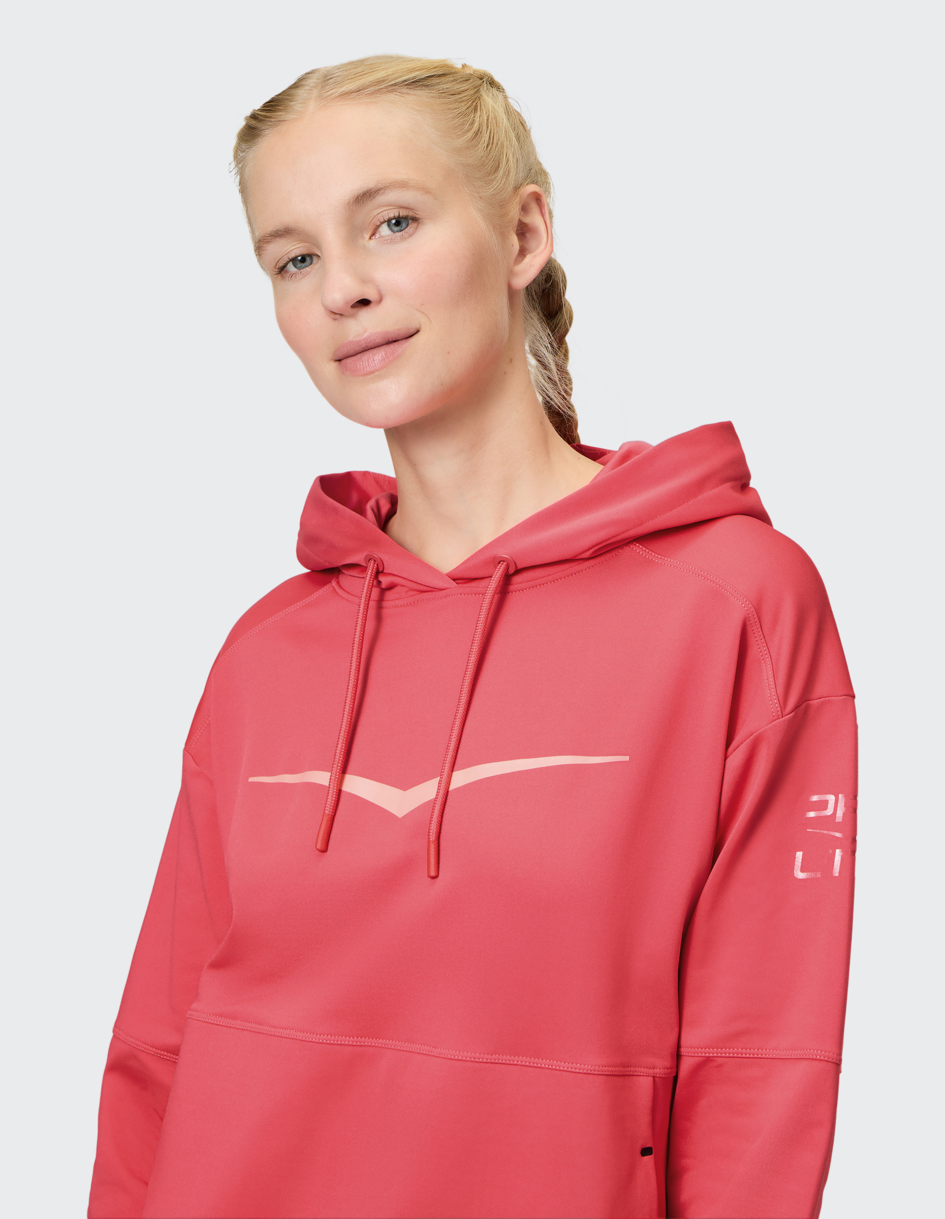 Eine Frau mit blondem, geflochtenem Haar trägt einen lockeren, roten Kapuzenpullover mit einem hellen Logo auf der Brust.