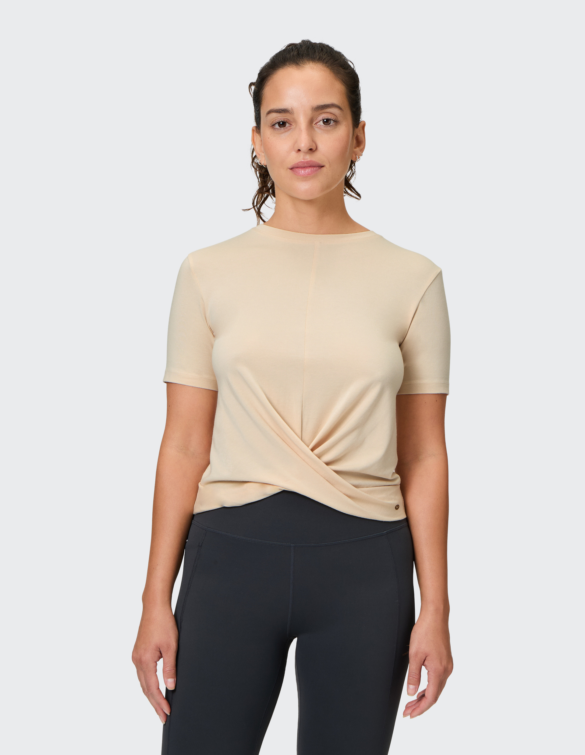 Eine Frau trägt ein beigefarbenes T-Shirt mit Twist-Detail vorne und schwarze Leggings, vor einem hellgrauen Hintergrund.