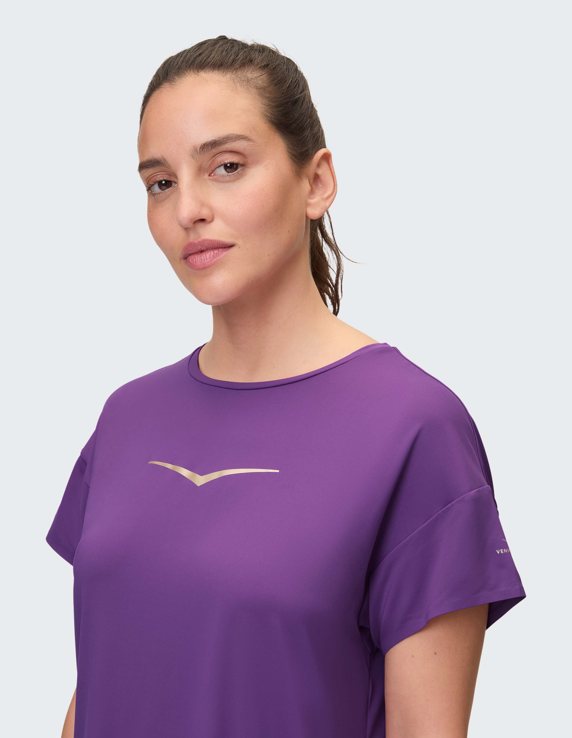 Frau mit zurückgebundenem Haar trägt ein lila T-Shirt mit V-Logo, schaut direkt in die Kamera, vor einem hellen Hintergrund.