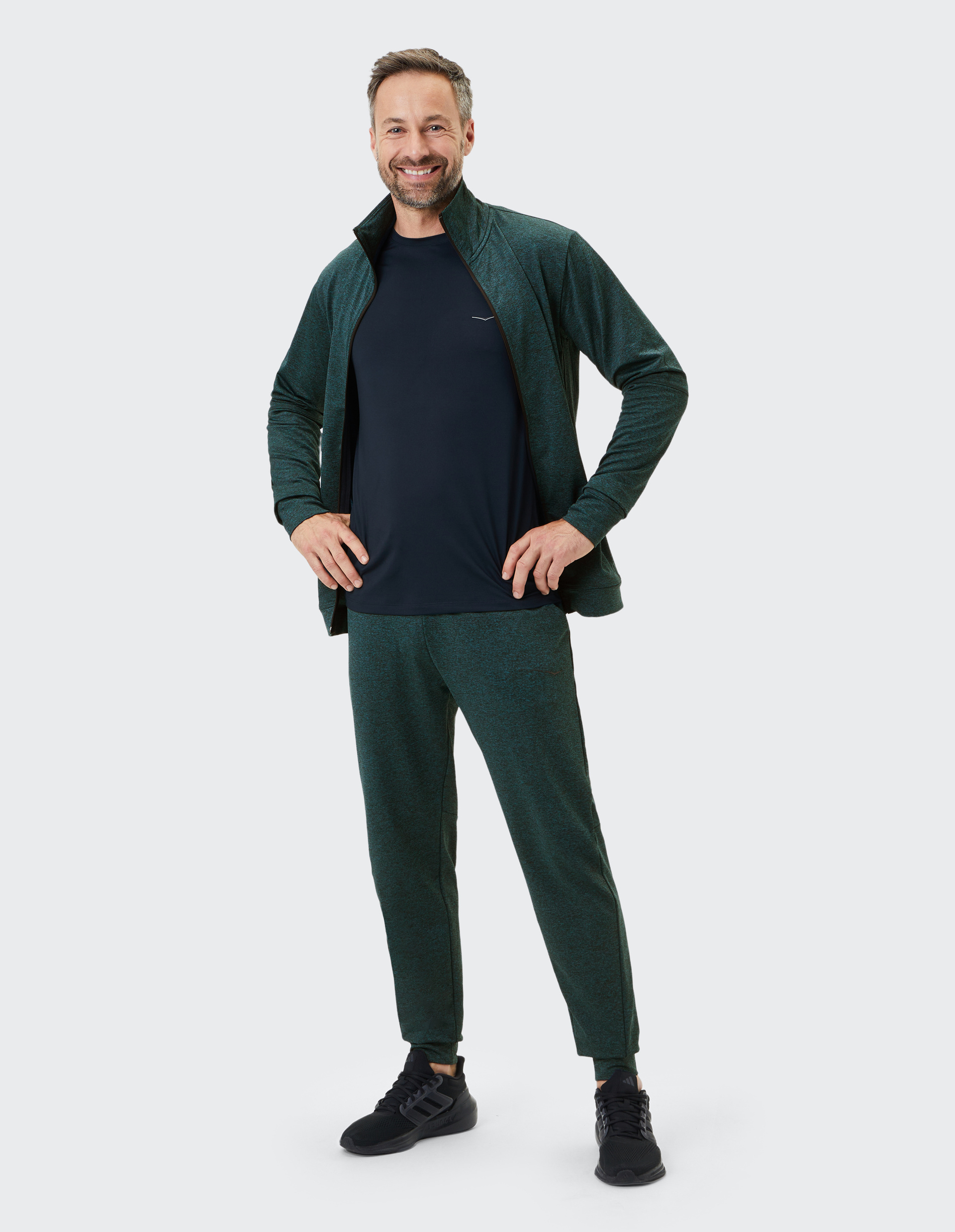 Ein Mann lächelt in sportlicher, dunkelgrüner Freizeitkleidung mit schwarzem Shirt, Händen in Hüften gestützt, auf einem hellen Hintergrund.