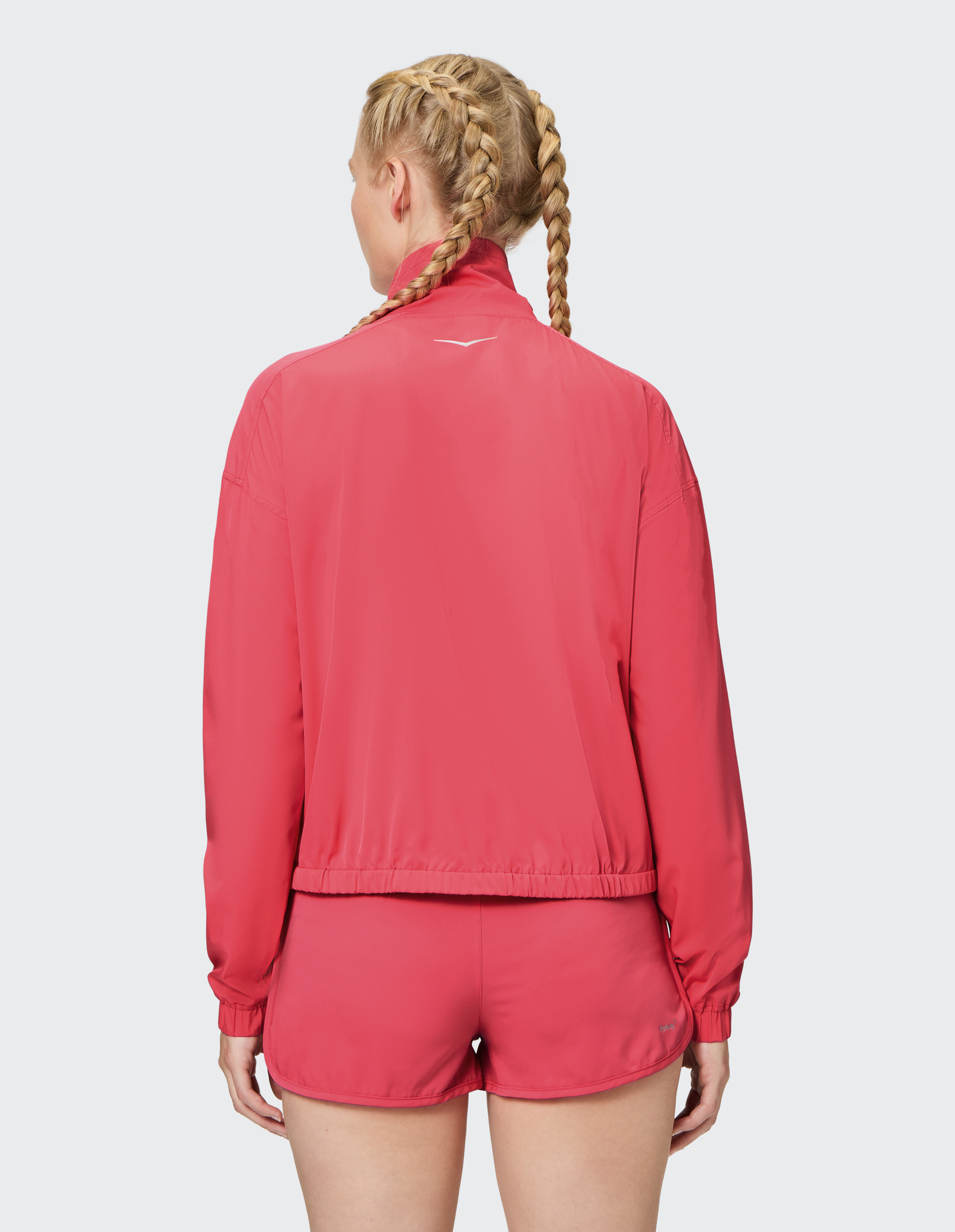Frau mit zwei geflochtenen Zöpfen trägt eine pinke Sportjacke und passende Shorts, steht mit dem Rücken zur Kamera.