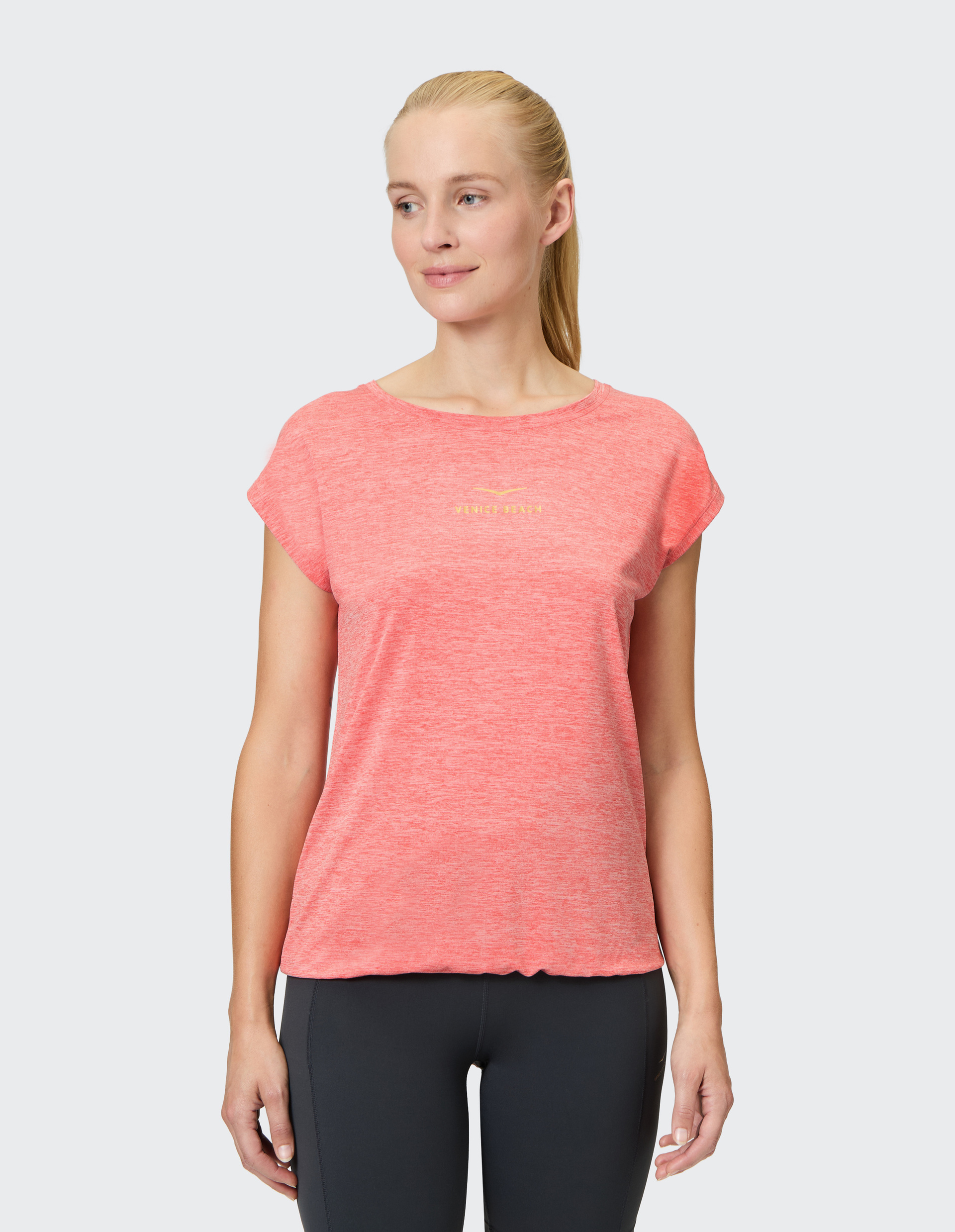 Frau in einem rosafarbenen T-Shirt steht lächelnd, mit langen Haaren zum Zopf gebunden, und trägt dunkle Leggings.