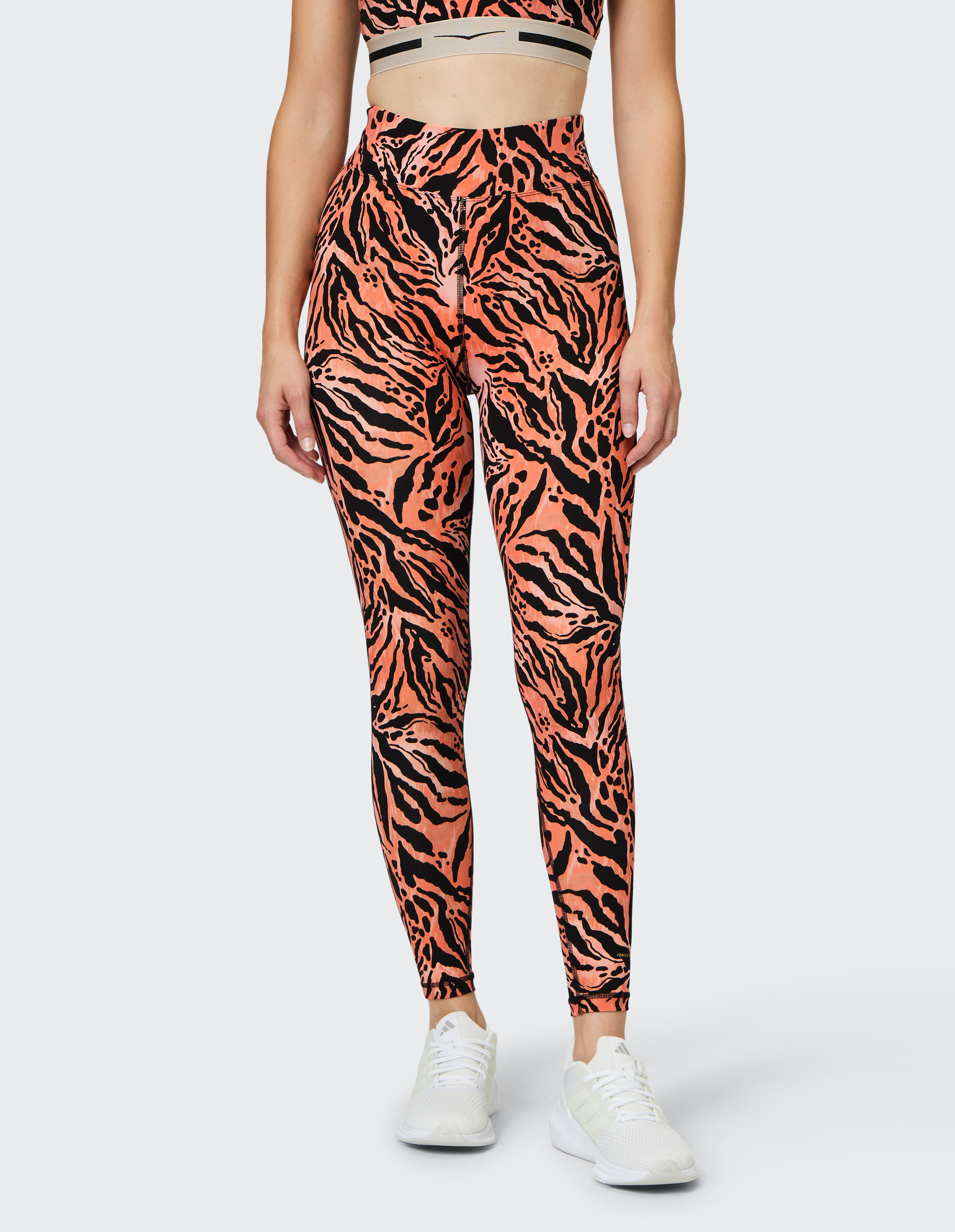 Frau trägt leggings mit orange-schwarzem Tigerstreifenmuster, dazu ein passendes kurzes Oberteil und weiße Sportschuhe.