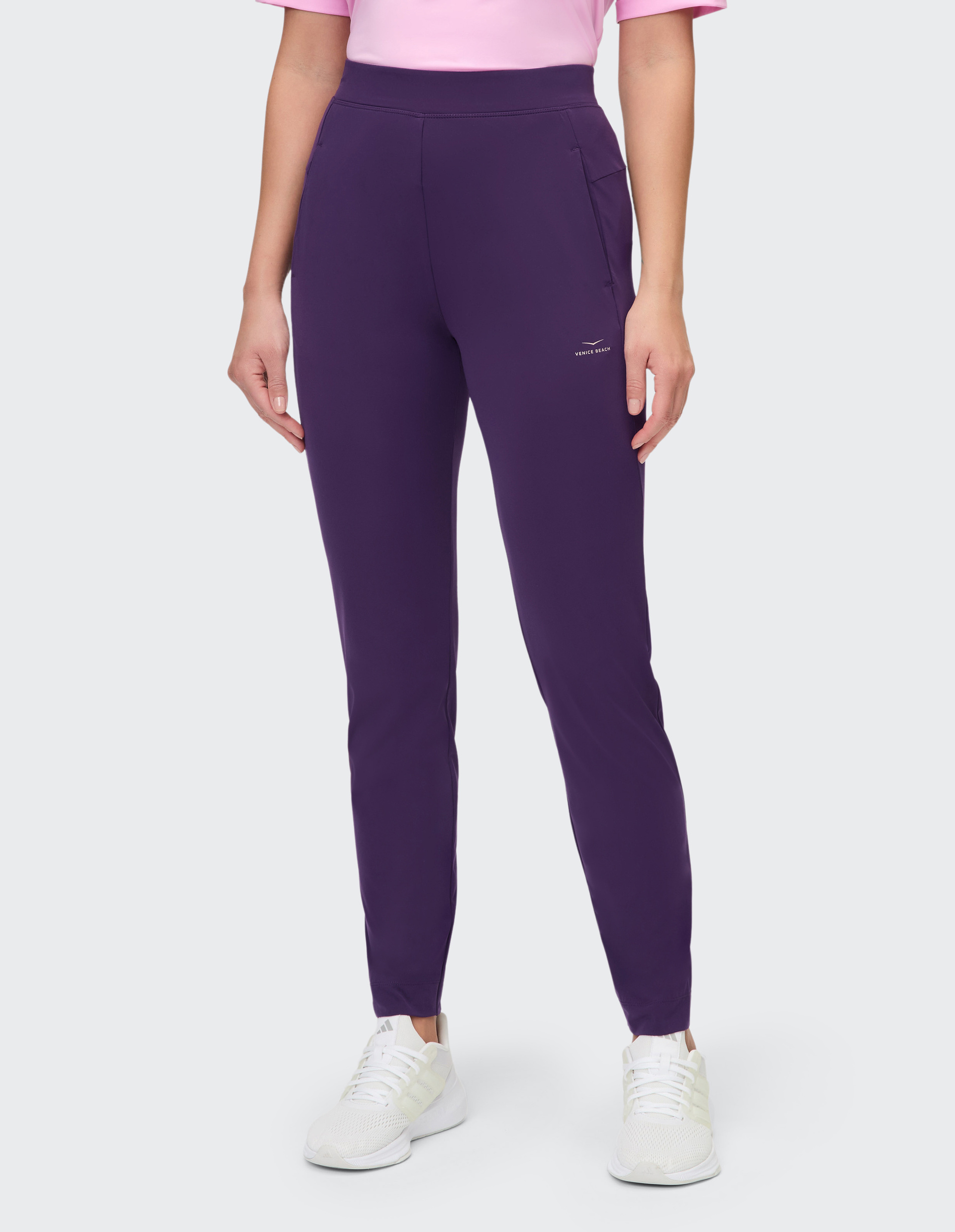 Frau in lila Sporthose und pinkem T-Shirt, trägt weiße Turnschuhe, vor grauem Hintergrund stehend.