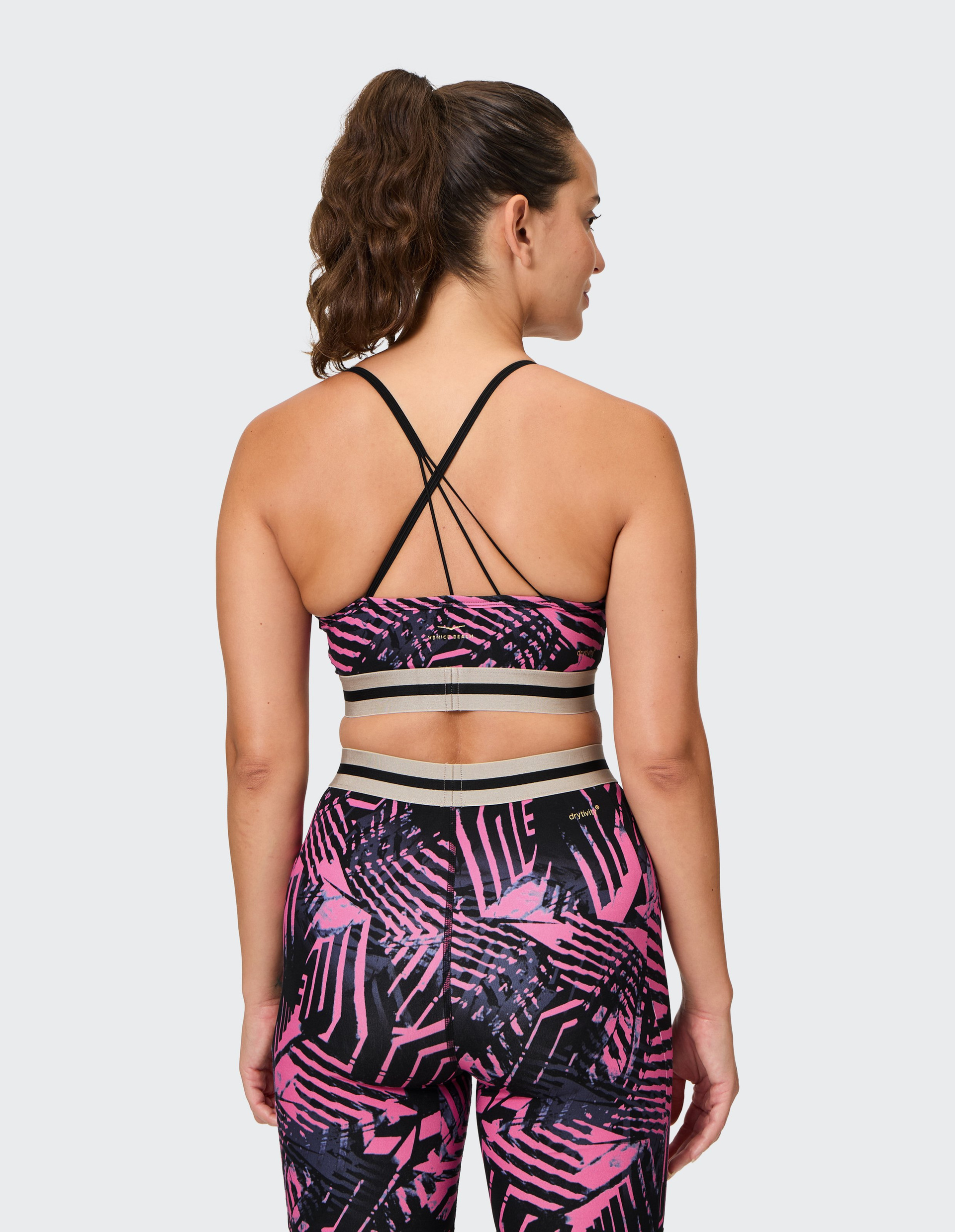 Frau von hinten in rosa-schwarz gemustertem Sport-Outfit mit gekreuzten Trägern, trägt Pferdeschwanz, vor grauem Hintergrund.