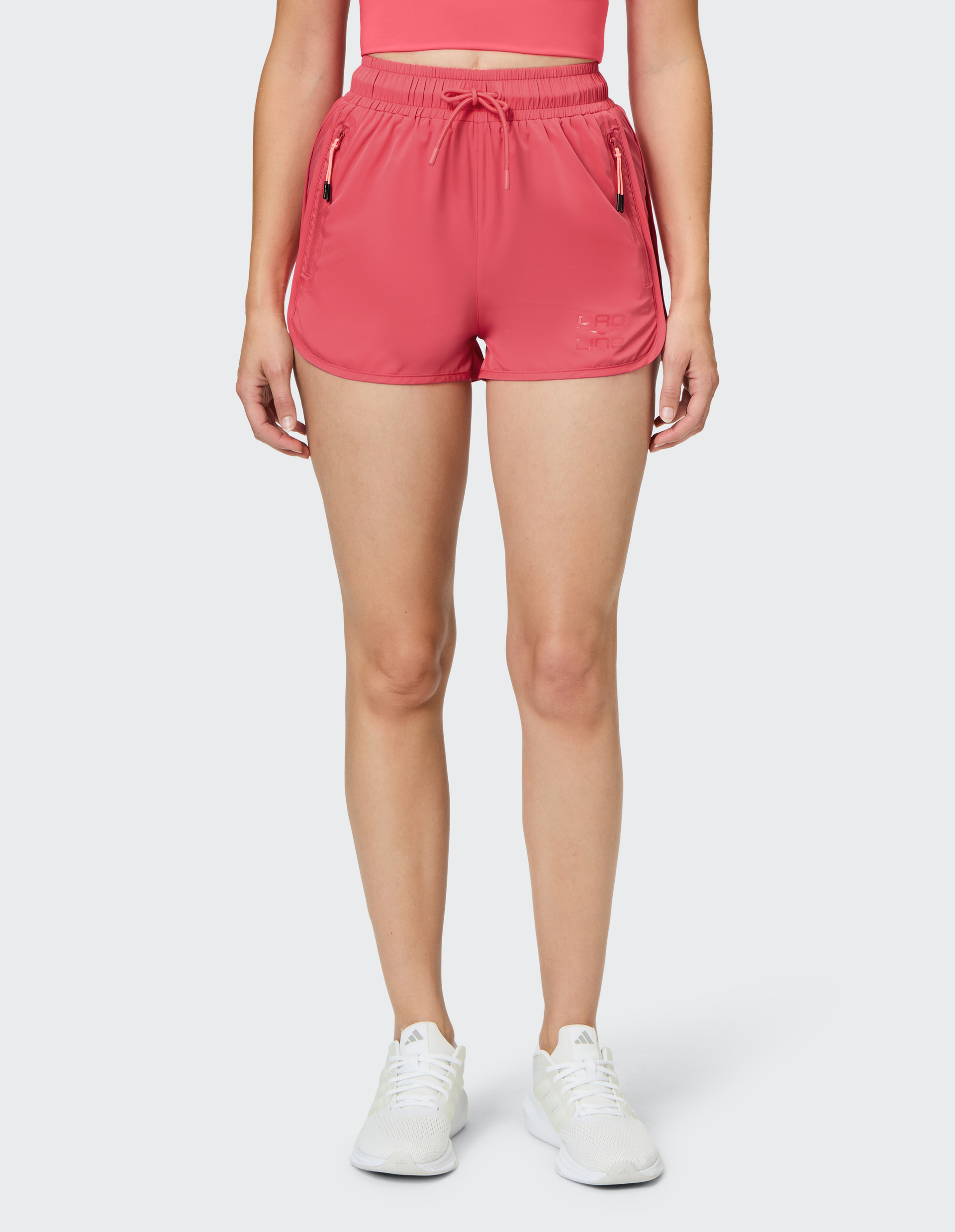 Frau trägt pinke Sportshorts mit Kordelzug und Reißverschlusstaschen, dazu ein pinkes Top und weiße Sportschuhe.