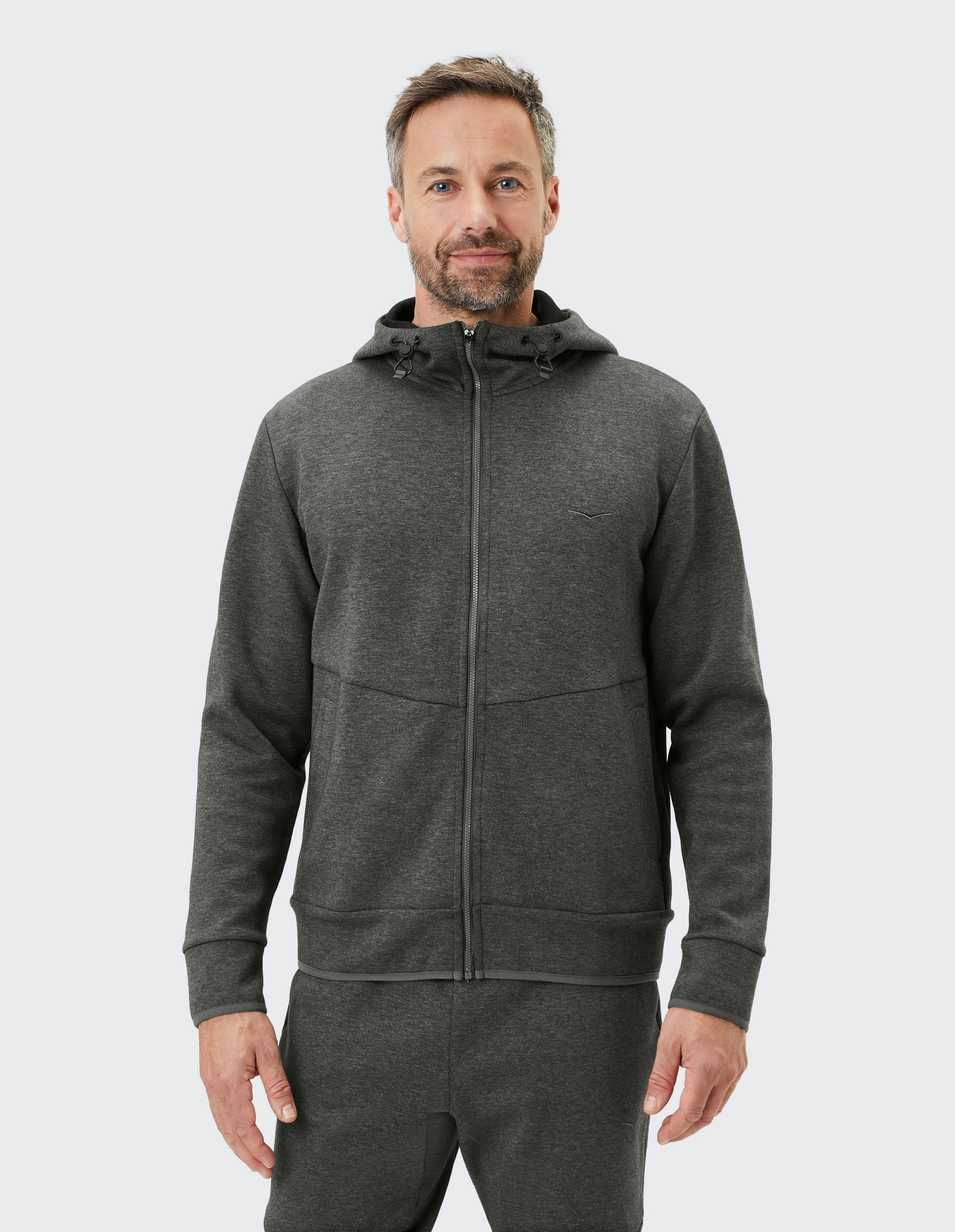 Ein Mann trägt einen dunkelgrauen Hoodie mit Reißverschluss und Kapuze sowie passende Jogginghosen, vor neutralem Hintergrund.
