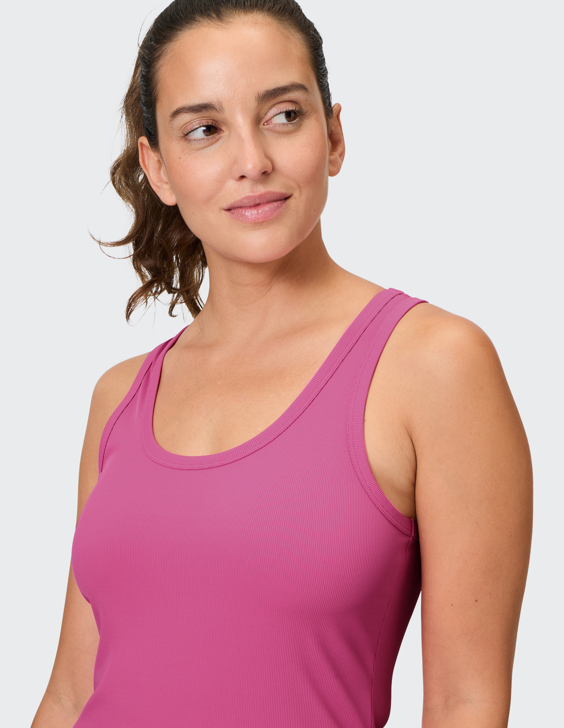 Eine Frau mit braunem Haar trägt ein pinkes Tanktop und blickt lächelnd zur Seite vor einem hellgrauen Hintergrund.