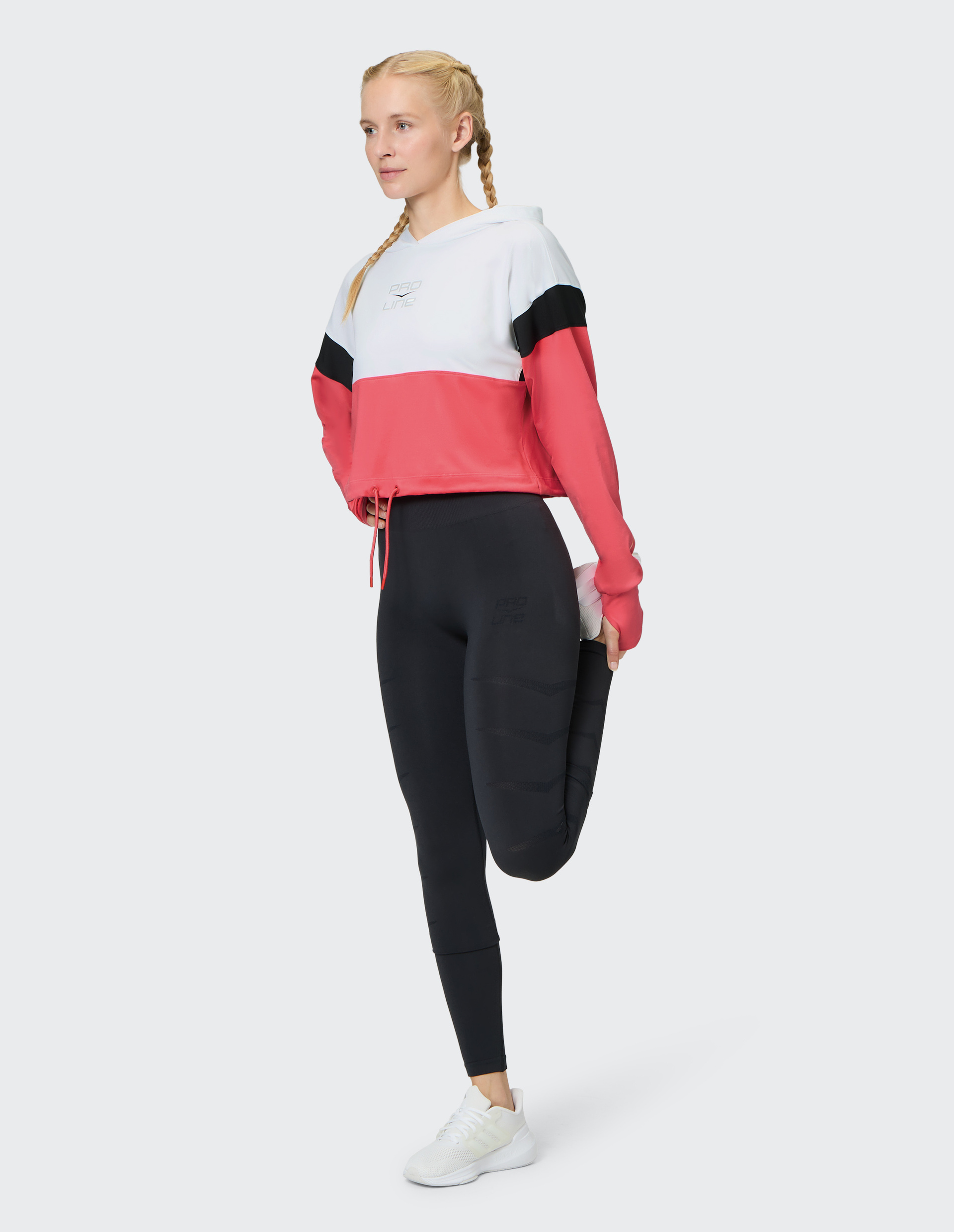 Eine Frau in einem rot-weißen Hoodie und schwarzen Leggings dehnt ihr Bein, während sie auf weißen Turnschuhen steht.