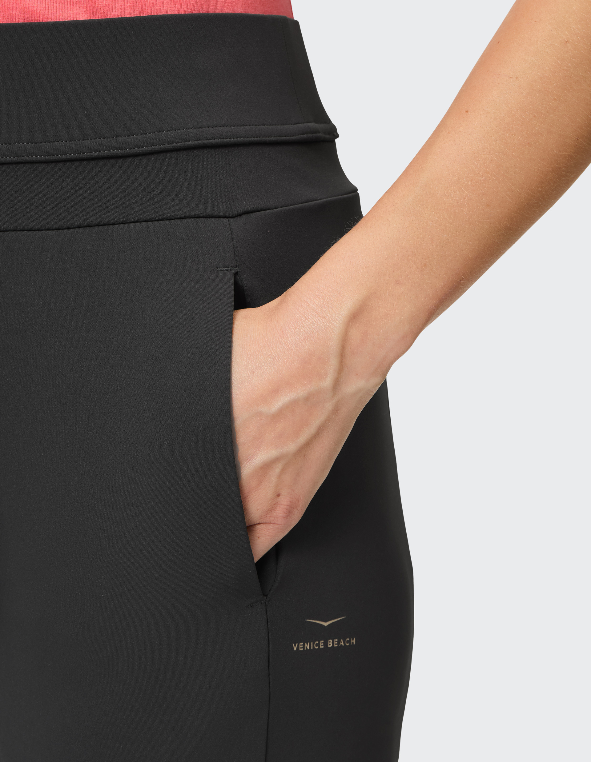 Nahaufnahme einer Hand in der Tasche einer schwarzen Venice Beach Sporthose, kombiniert mit einem roten Oberteil.