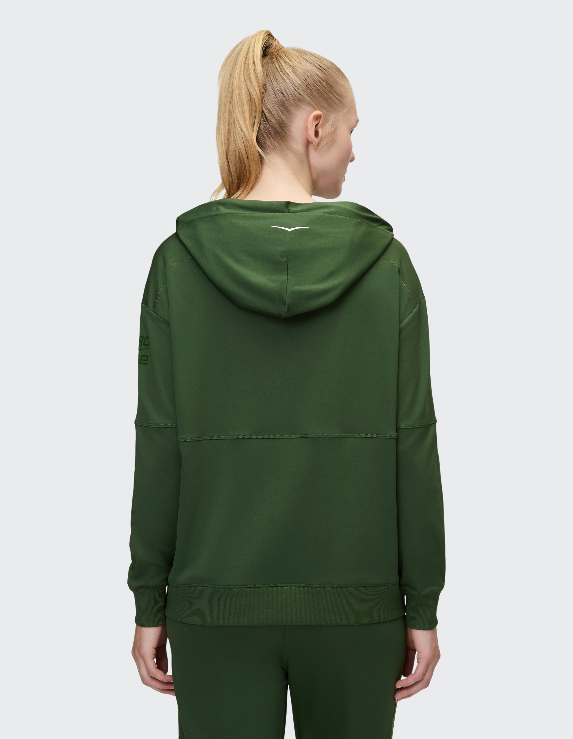 Person mit blondem Pferdeschwanz trägt von hinten gesehen einen dunkelgrünen Hoodie und passende Hose vor grauem Hintergrund.