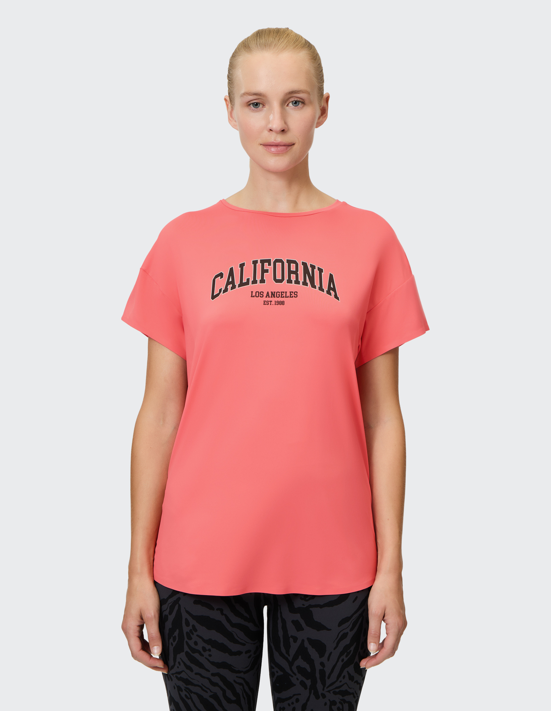 Frau trägt ein pinkes T-Shirt mit "California Los Angeles" Aufdruck und schwarze gemusterte Hose, vor grauem Hintergrund stehend.