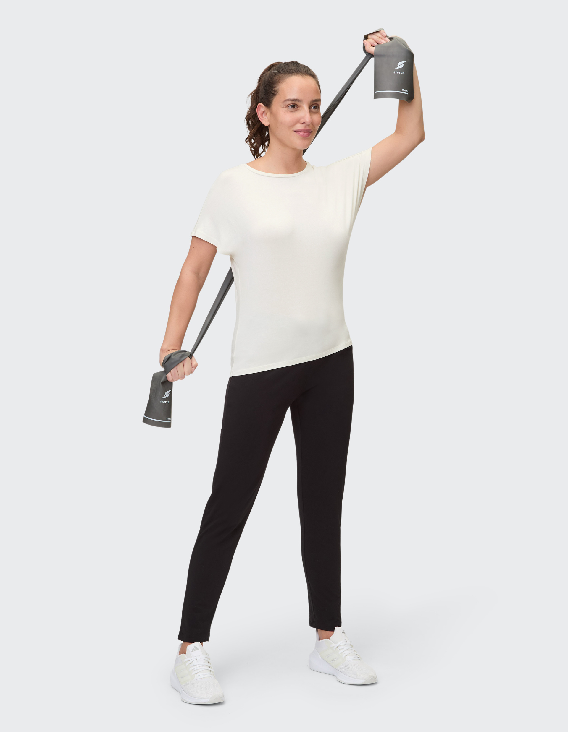 Eine Frau trägt ein weißes T-Shirt und schwarze Hosen, steht frontal auf hellem Hintergrund und trägt weiße Turnschuhe.