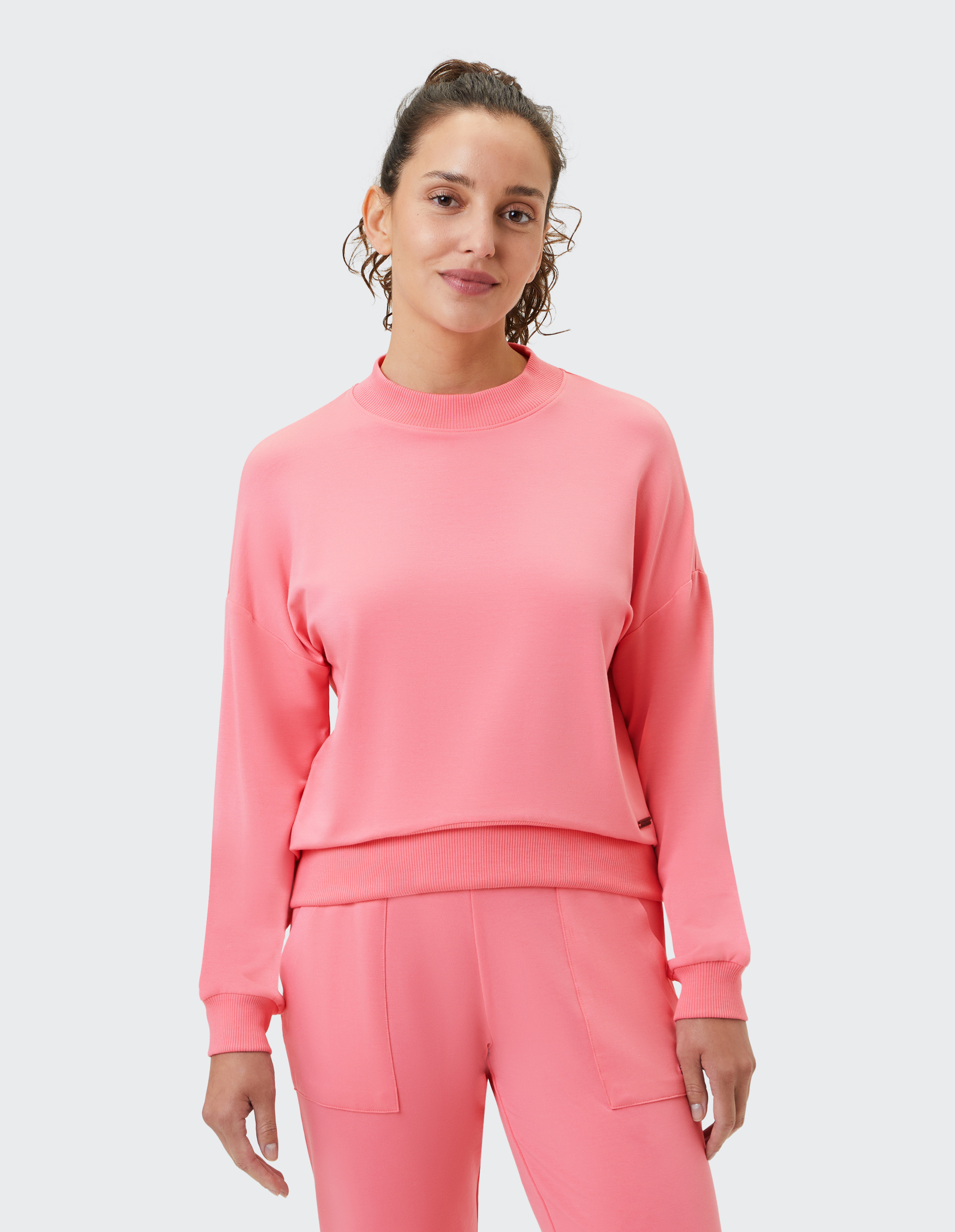 Frau in einem korallfarbenen Sweatshirt und passender Hose lächelt leicht, vor weißem Hintergrund stehend.