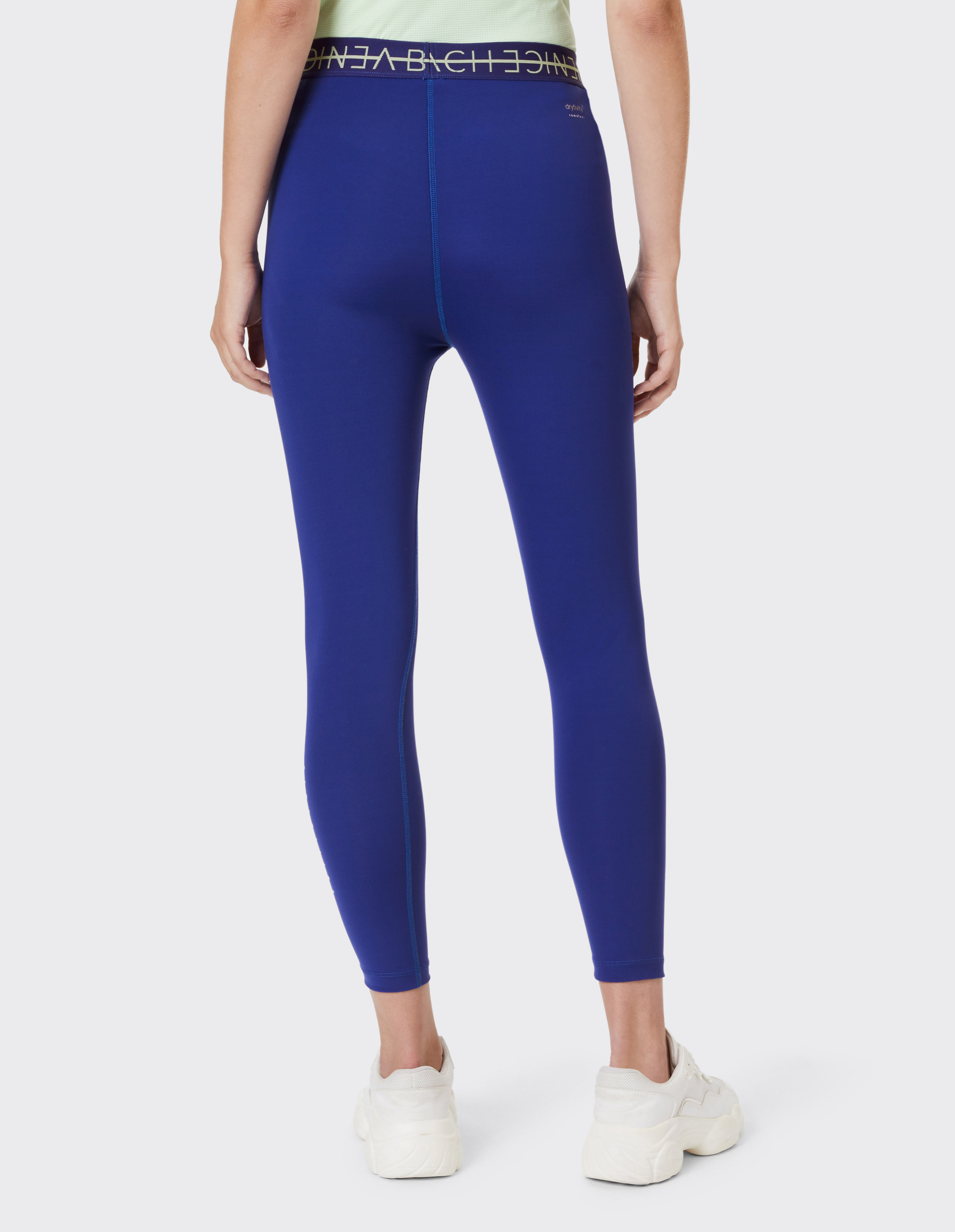 Person trägt eine blaue Leggings mit elastischem Bund und weißen Turnschuhen, fotografiert von hinten, auf hellgrauem Hintergrund.