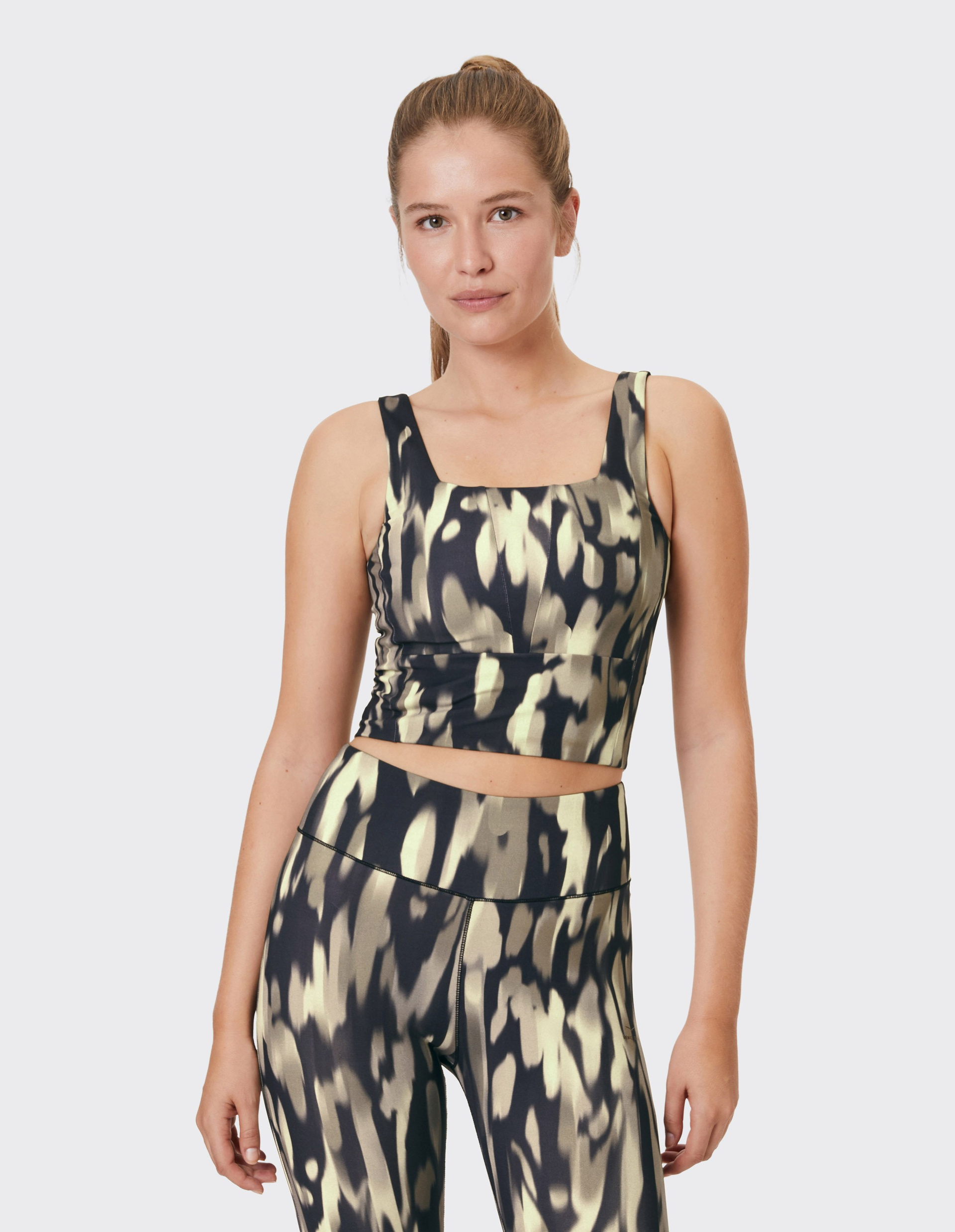 Junge Frau trägt ein figurbetontes, gemustertes Sport-Top mit passenden Leggings vor neutralem Hintergrund.