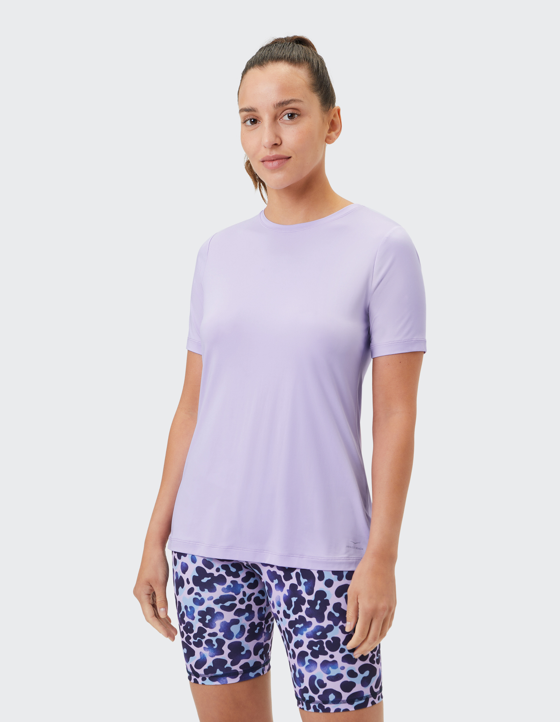 Frau in lavendelfarbenem T-Shirt und gemusterten blauen Shorts mit Leopardenmuster vor weißem Hintergrund.