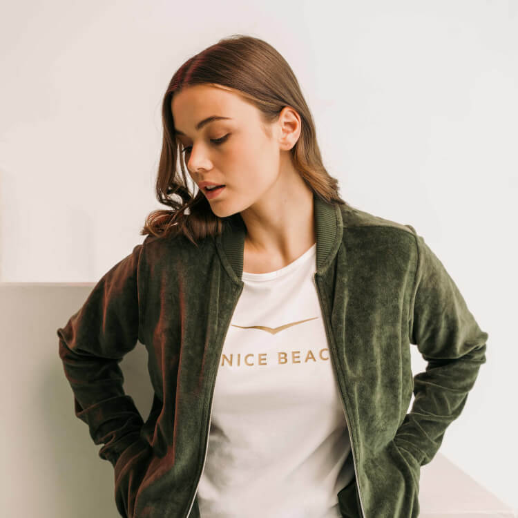 Frau in dunkelgrüner Jacke und weißem T-Shirt mit goldenem Venice Beach Logo.